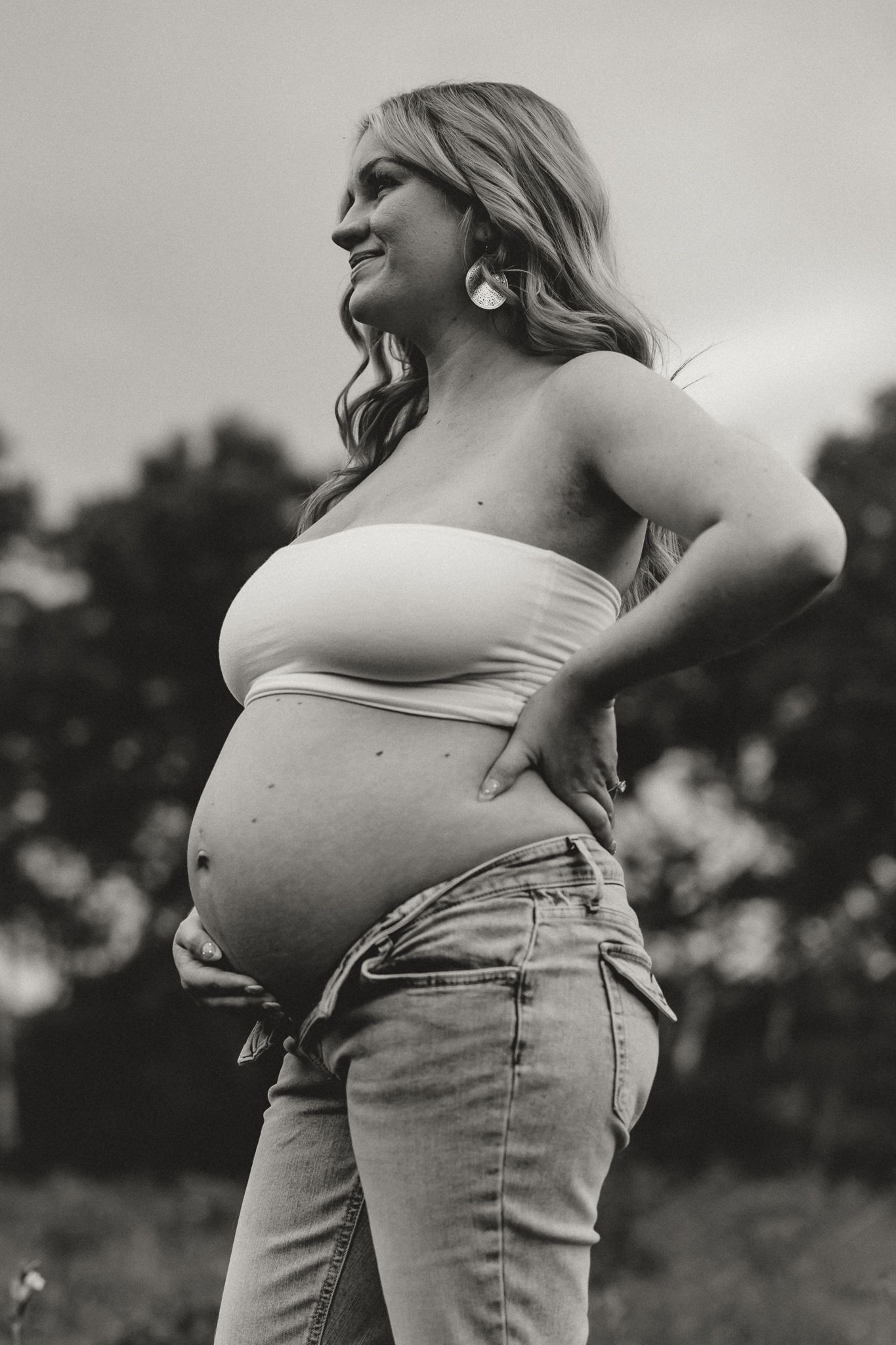Black and white maternity photo of pregnant blonde woman in white tube top and jeans outdoors.