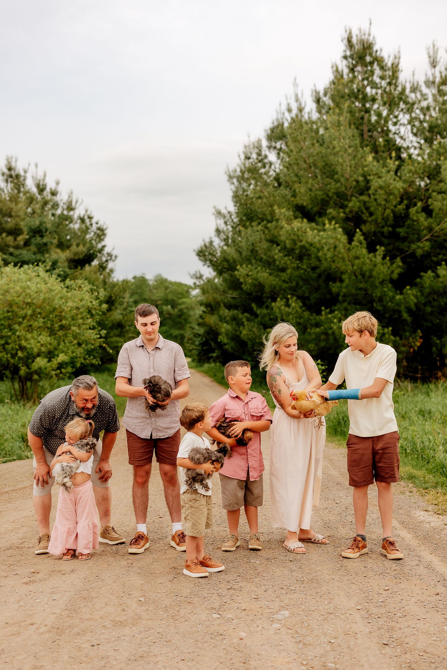 Extended family holding chickens on a dirt path surrounded by green trees on a cloudy day.