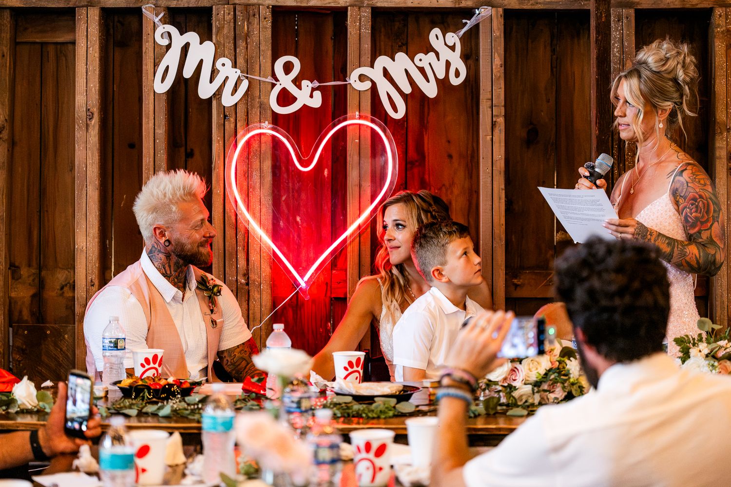 Couple at wedding reception table with neon heart and Mr & Mrs sign, while a guest gives a speech.
