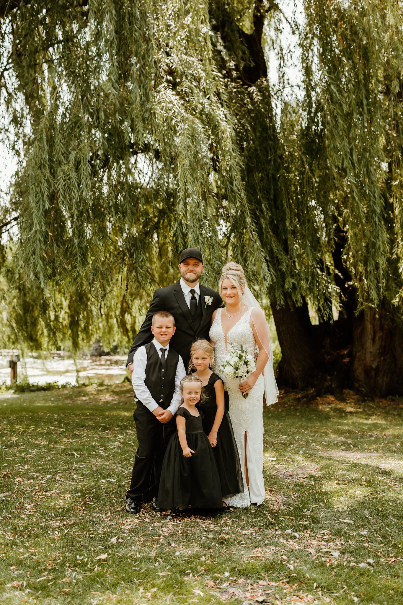 Bride and groom pose with two children under a weeping willow tree, bride holding white bouquet in elegant white gown.