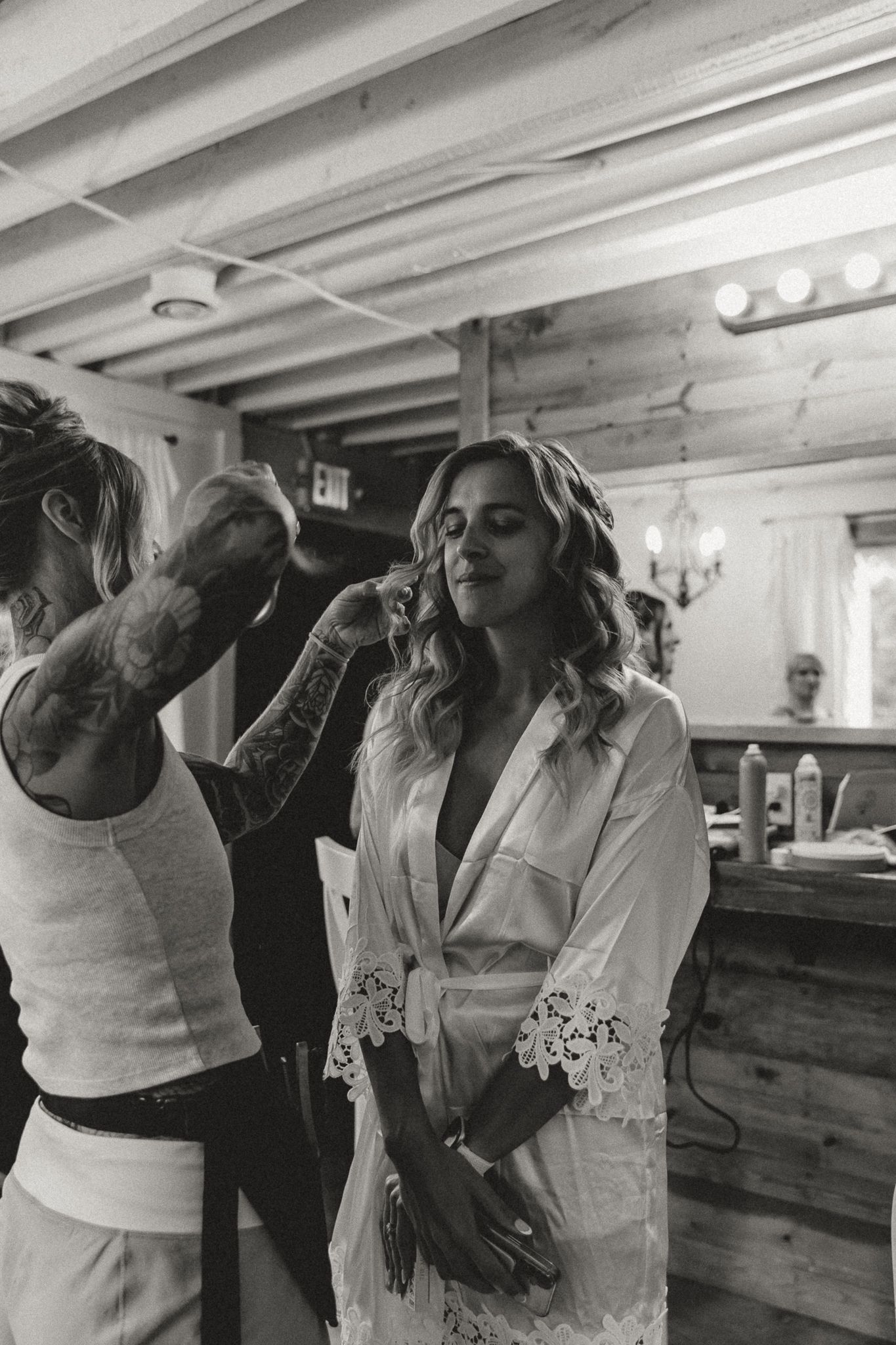 Black and white photo of a bride in a lace robe getting hair and makeup done by a tattooed stylist in a rustic venue.