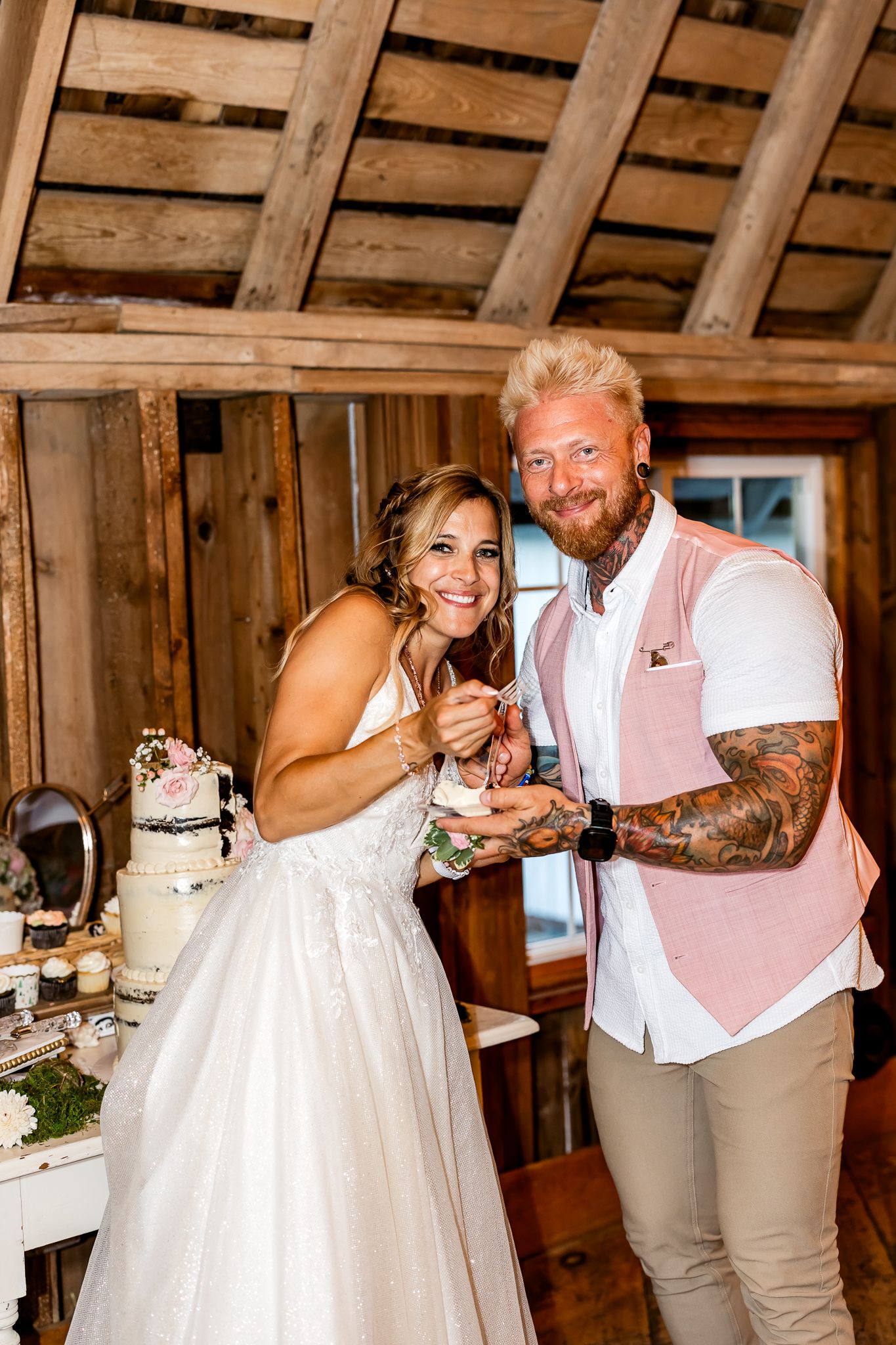 Bride and groom smiling while cutting wedding cake in a rustic barn venue, groom wearing pink vest with tattoos.