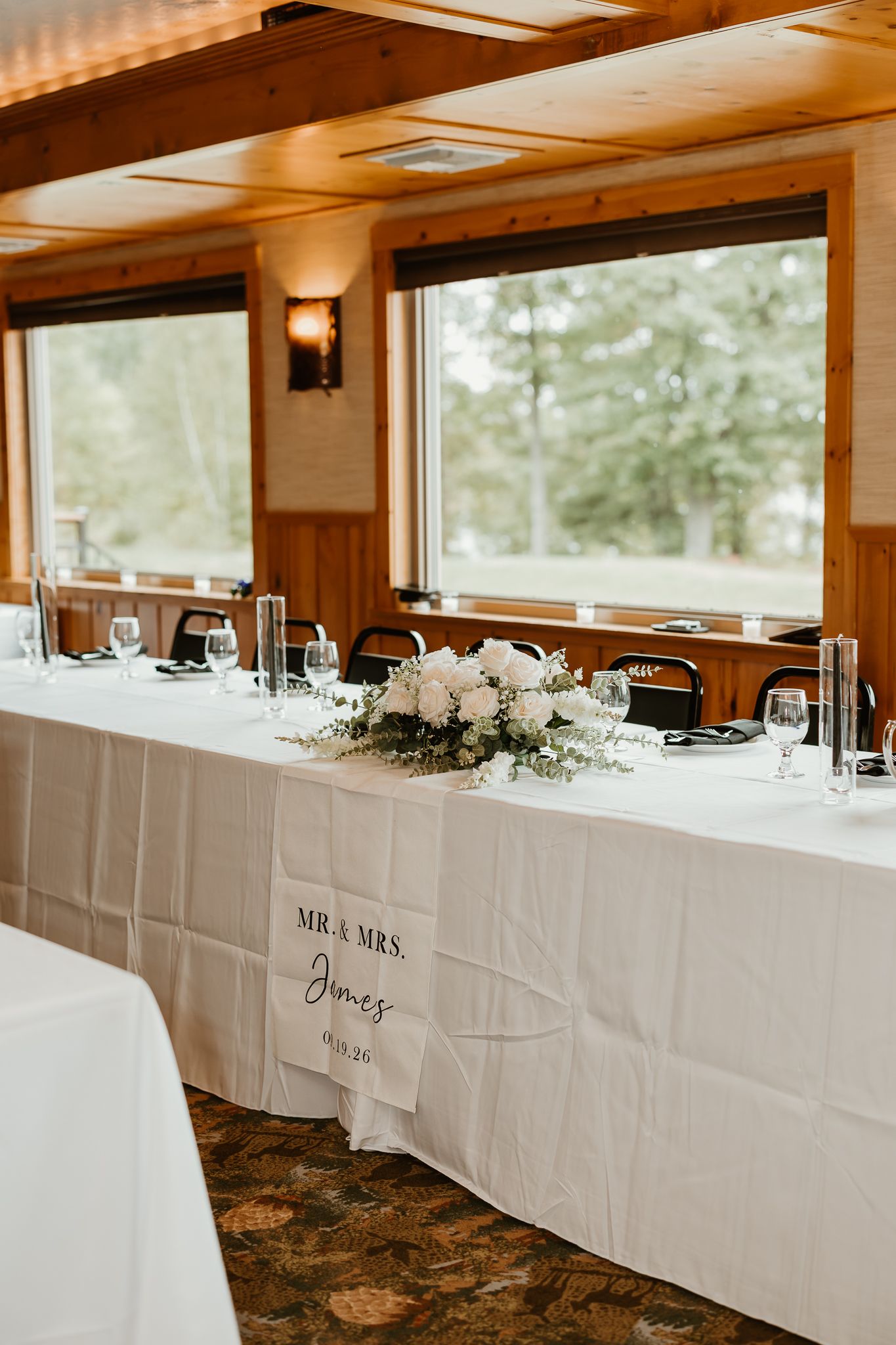 Wedding head table with white linens, floral centerpiece and a Mr. and Mrs. Jones banner, set beside large windows overlooking green trees.