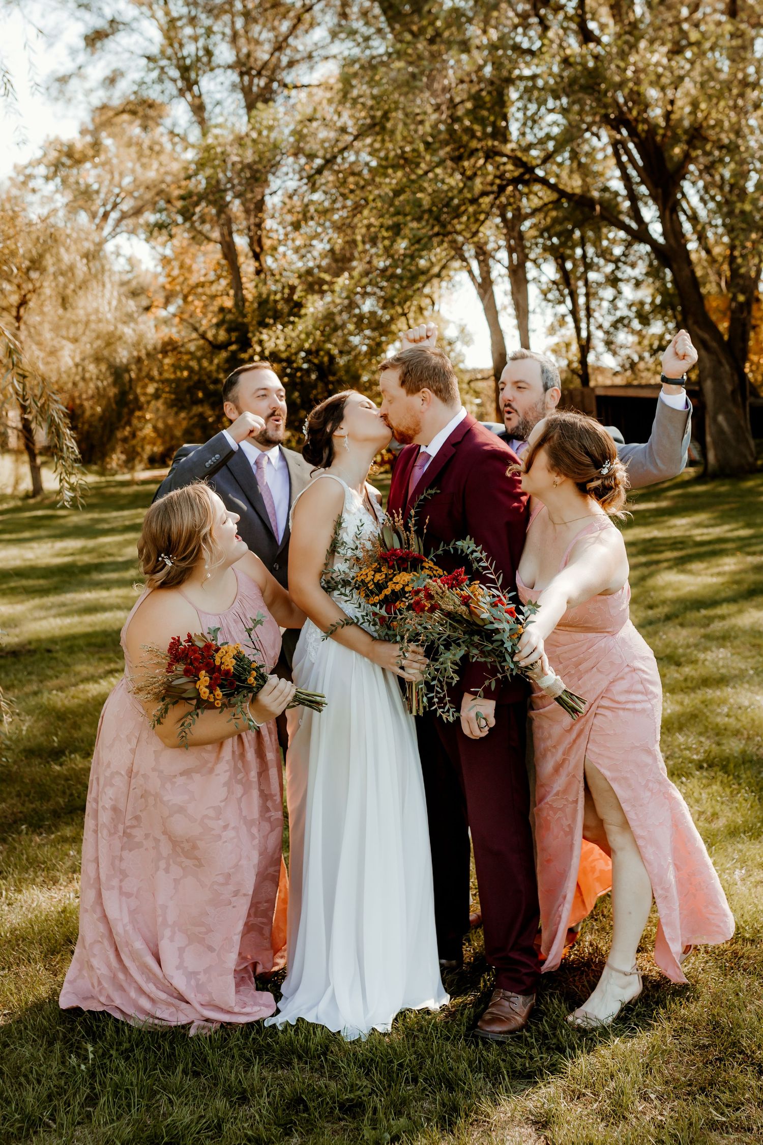 Bride in a white gown kisses the groom in a burgundy suit as bridesmaids in blush dresses and groomsmen cheer around them in a sunny park.