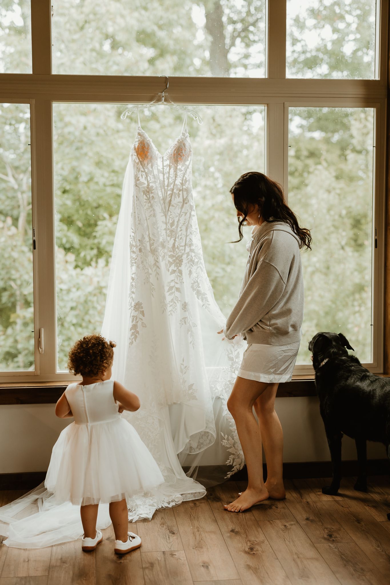 Bride in a gray sweatshirt and a flower girl in a white tutu dress admire a lace wedding gown hanging by a window as a black dog watches.