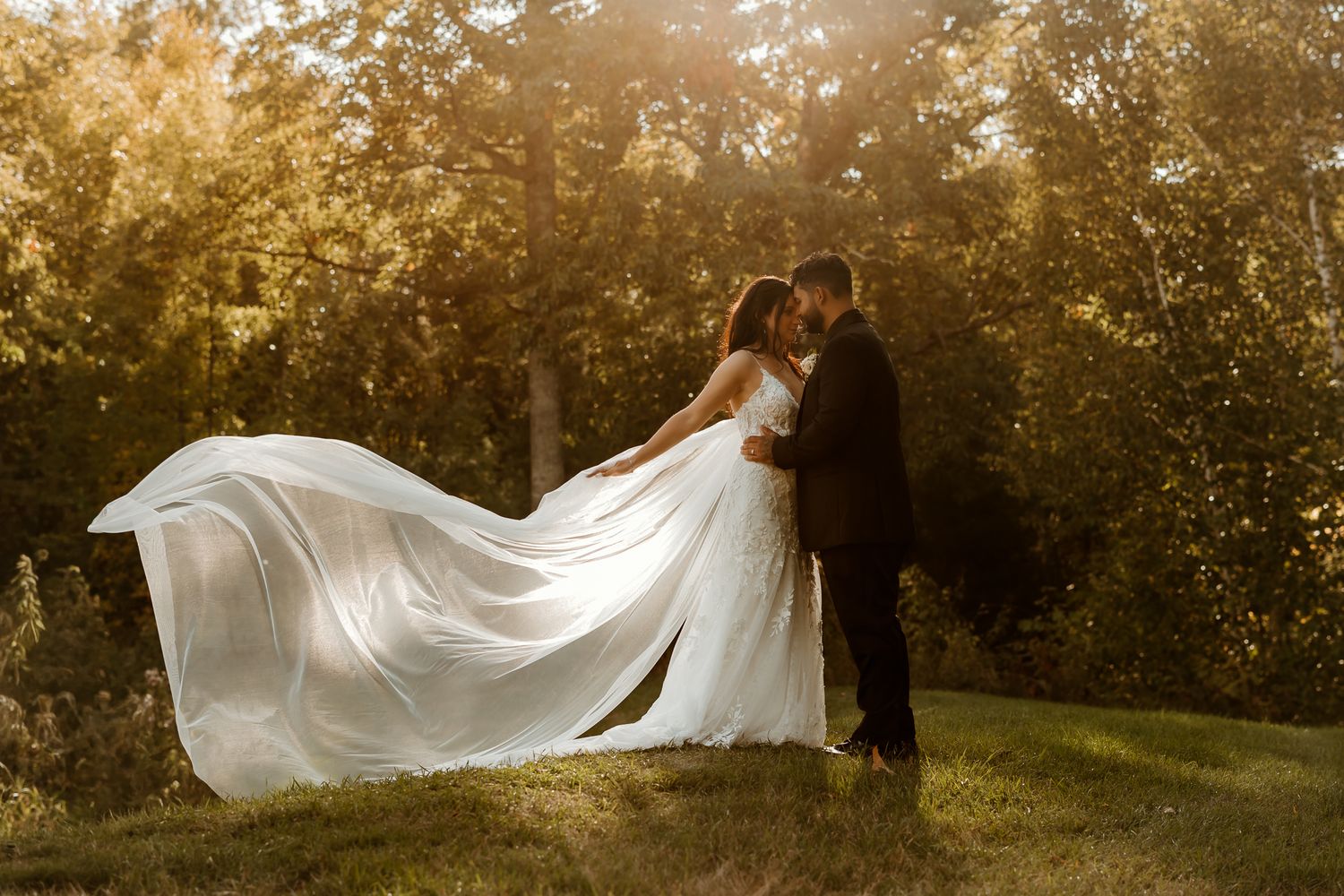 Bride in a flowing white gown with a long sheer train embraces her groom in a dark suit on a sunlit grassy hill surrounded by golden trees.