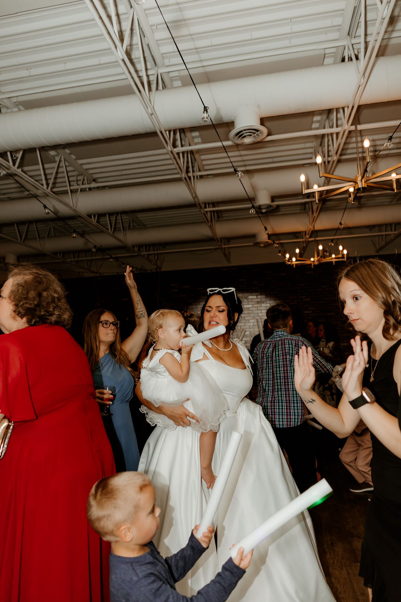 A bride in a white gown holds a toddler while guests clap and a young boy waves a rolled paper at a lively wedding reception.