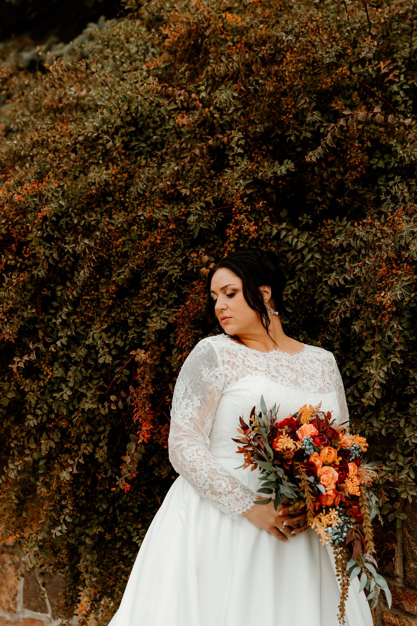 Bride in a white lace long-sleeve wedding gown holds an orange and red autumn bouquet while posing before a leafy, russet-colored hedge.