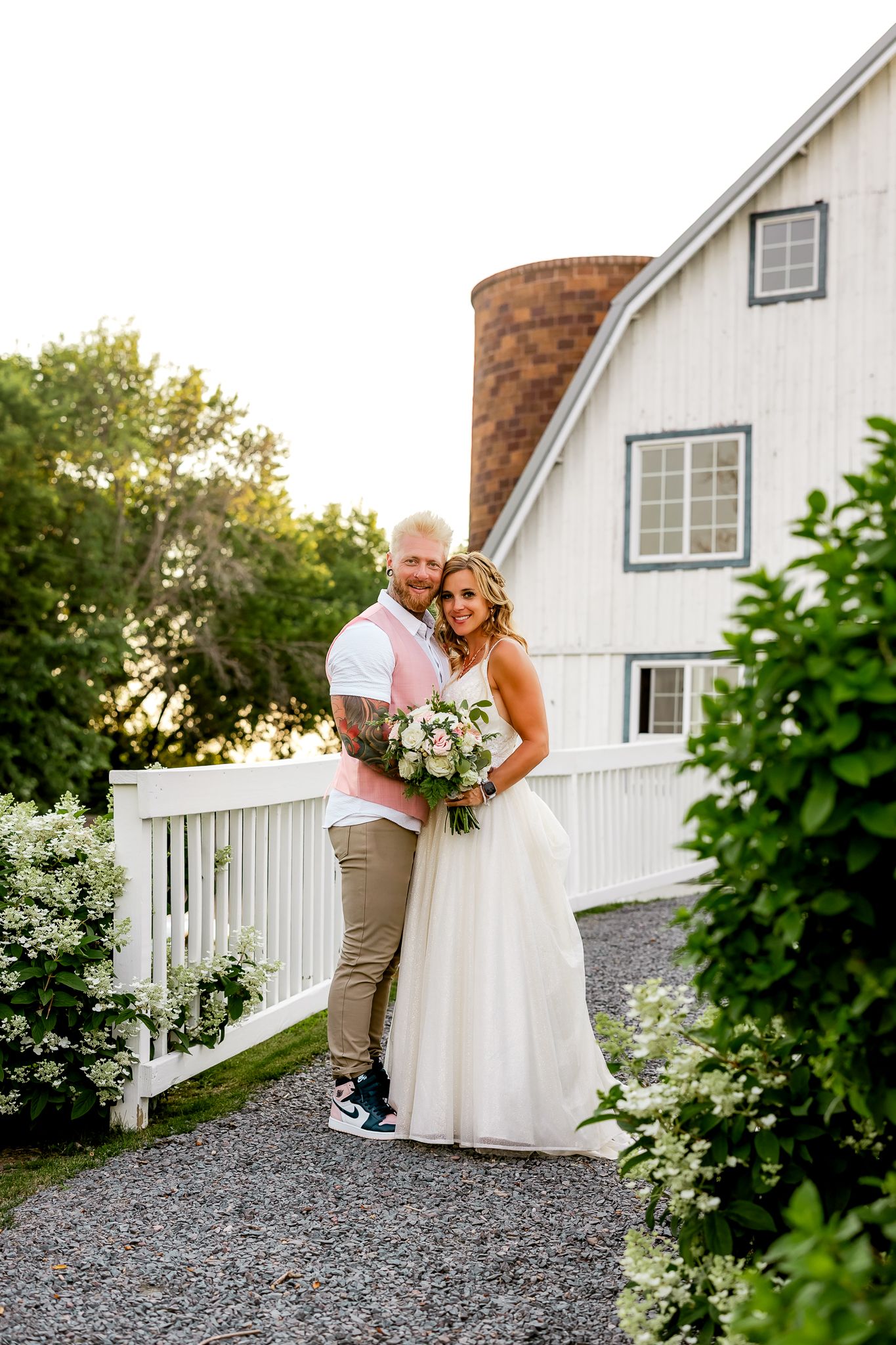 Bride and groom pose together outside a white barn venue, bride holding a floral bouquet on their wedding day.