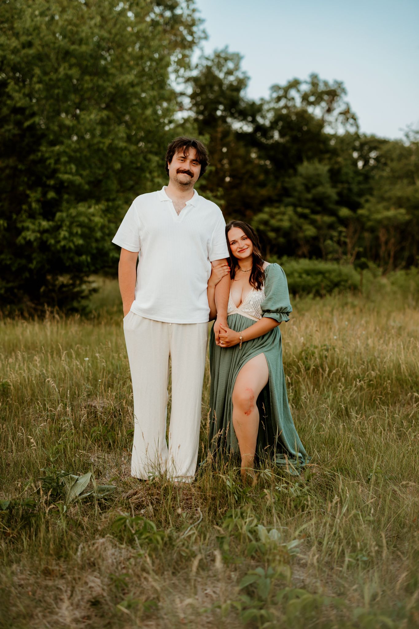 Couple posing in a green meadow, man in white outfit, woman in sage green maternity gown, surrounded by lush trees.