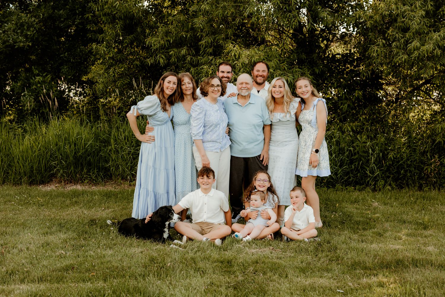 Large family group portrait outdoors on green grass, wearing coordinated light blue outfits, with trees in background.
