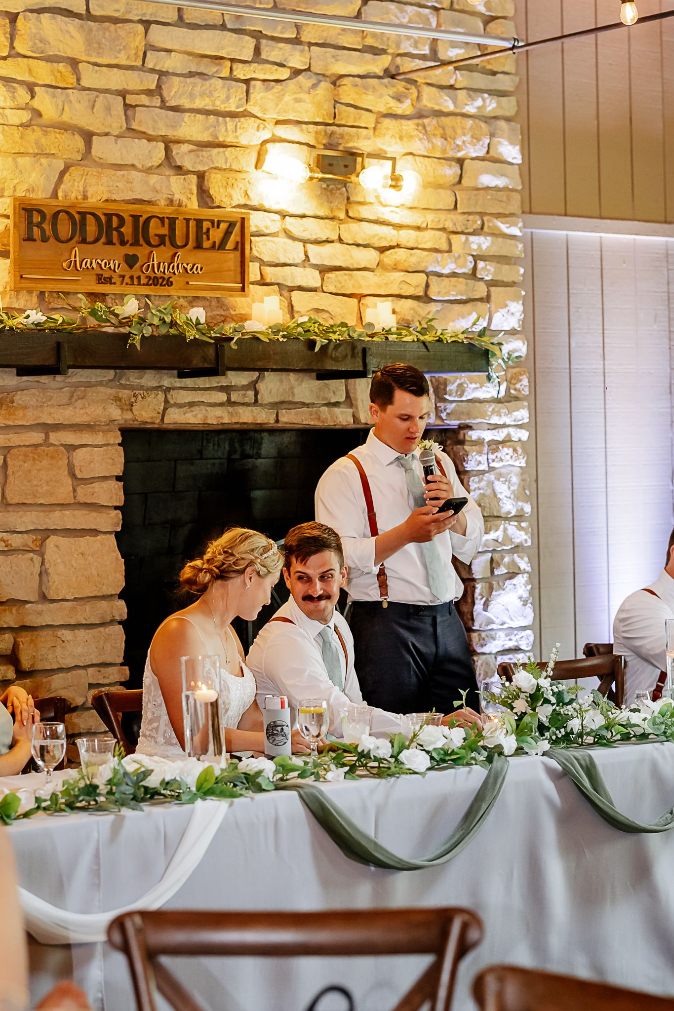Wedding reception toast at Rodriguez marriage celebration, groom reading speech near stone fireplace with floral table arrangements.