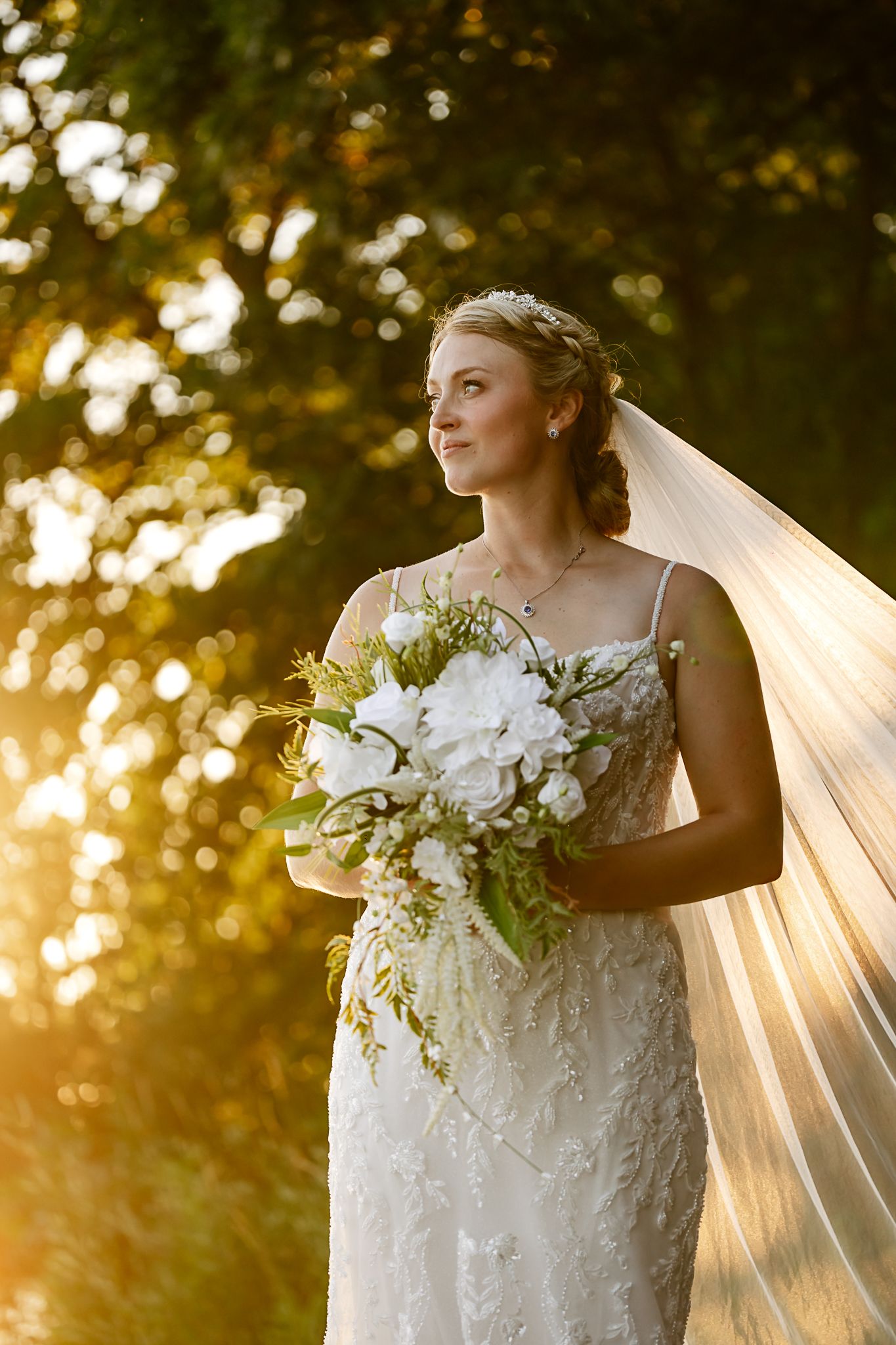 Bride in lace gown holding white floral bouquet with veil flowing in golden sunset light outdoors.