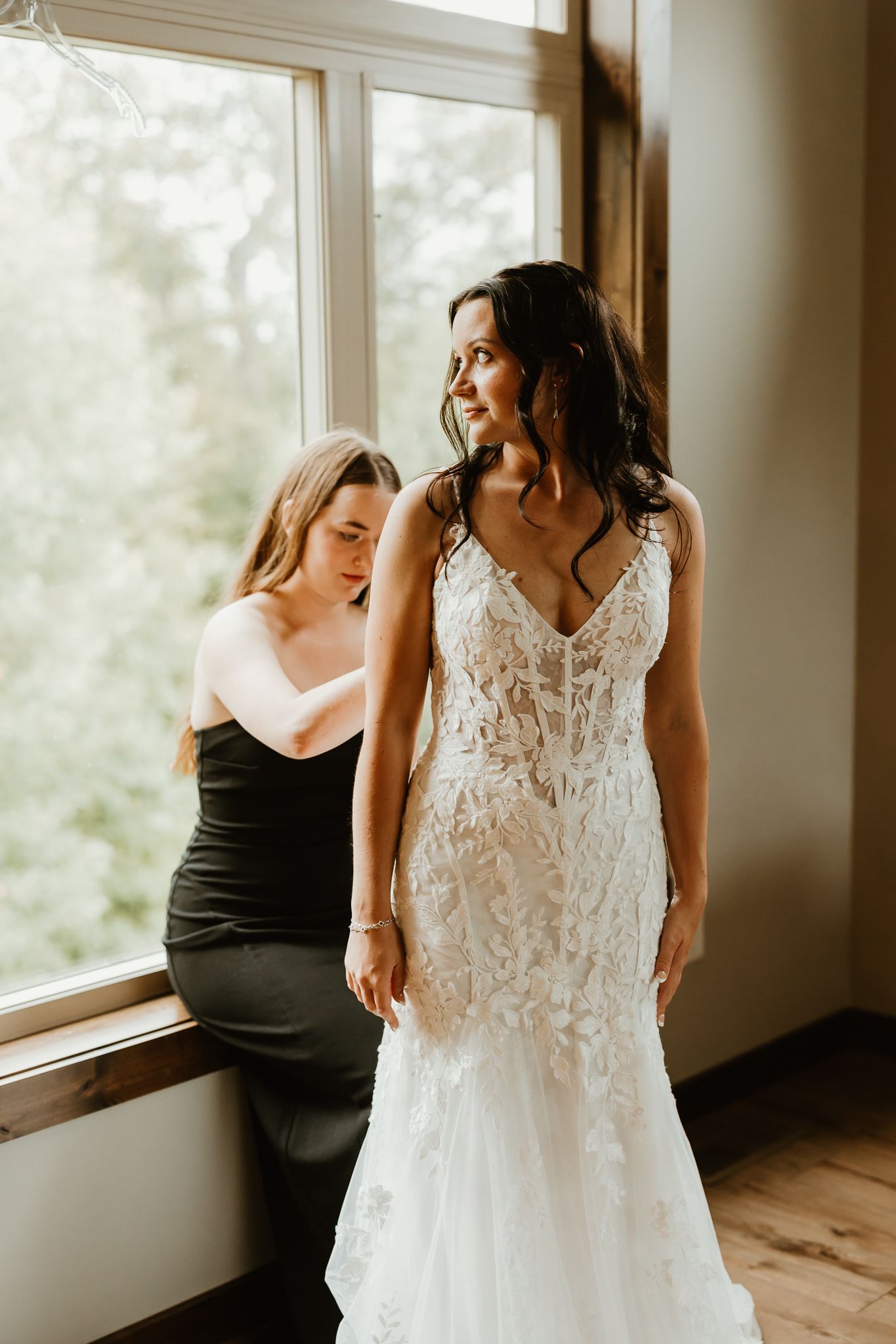 Bride in a white lace mermaid wedding gown stands by a sunlit window while a bridesmaid in black sits beside her.
