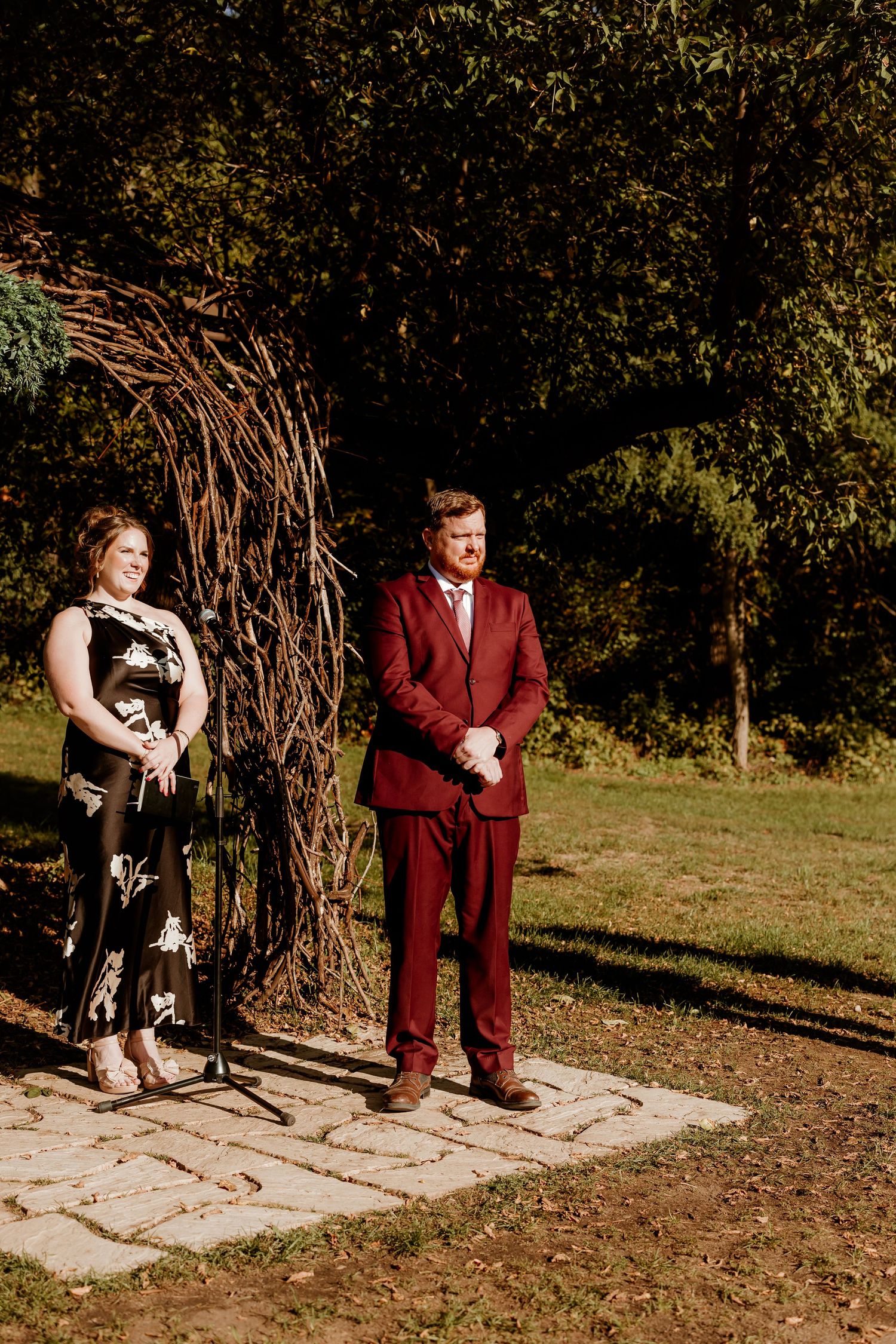 Man in a burgundy suit and woman in a black floral dress stand on a stone patio beside a woven branch arch at an outdoor wedding ceremony.