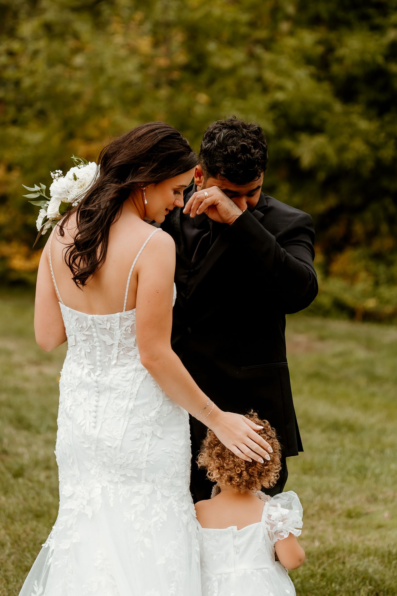 A bride in a lace gown with a floral hairpiece reaches toward a groom wiping his tears as a little girl in white looks on outdoors.