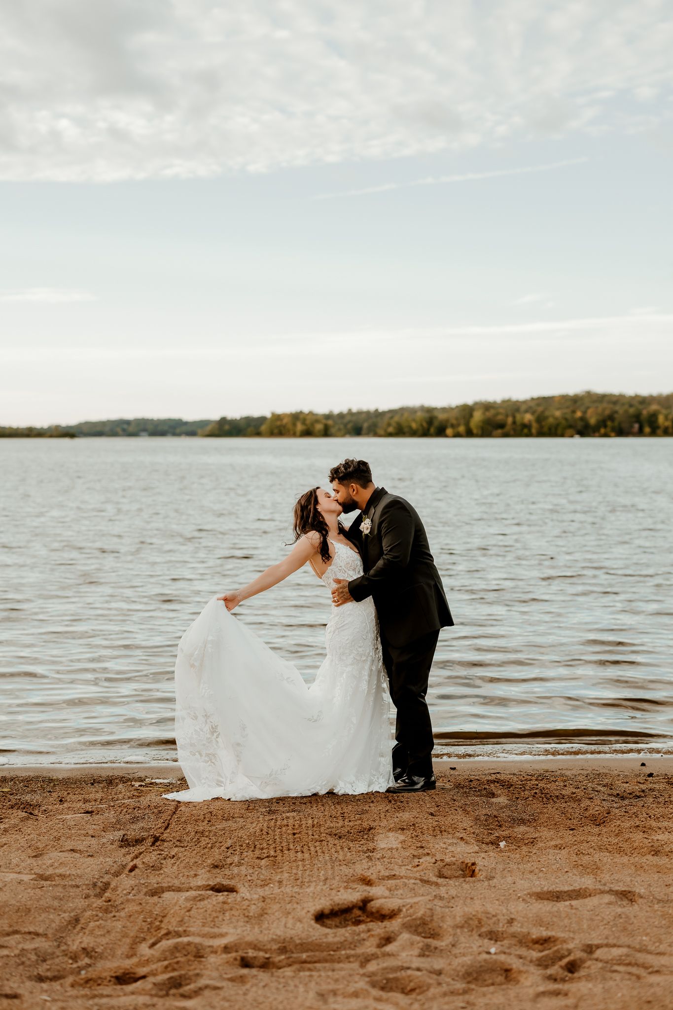 Bride in a flowing white lace gown kisses her groom in a dark suit on a sandy lakeshore at sunset.