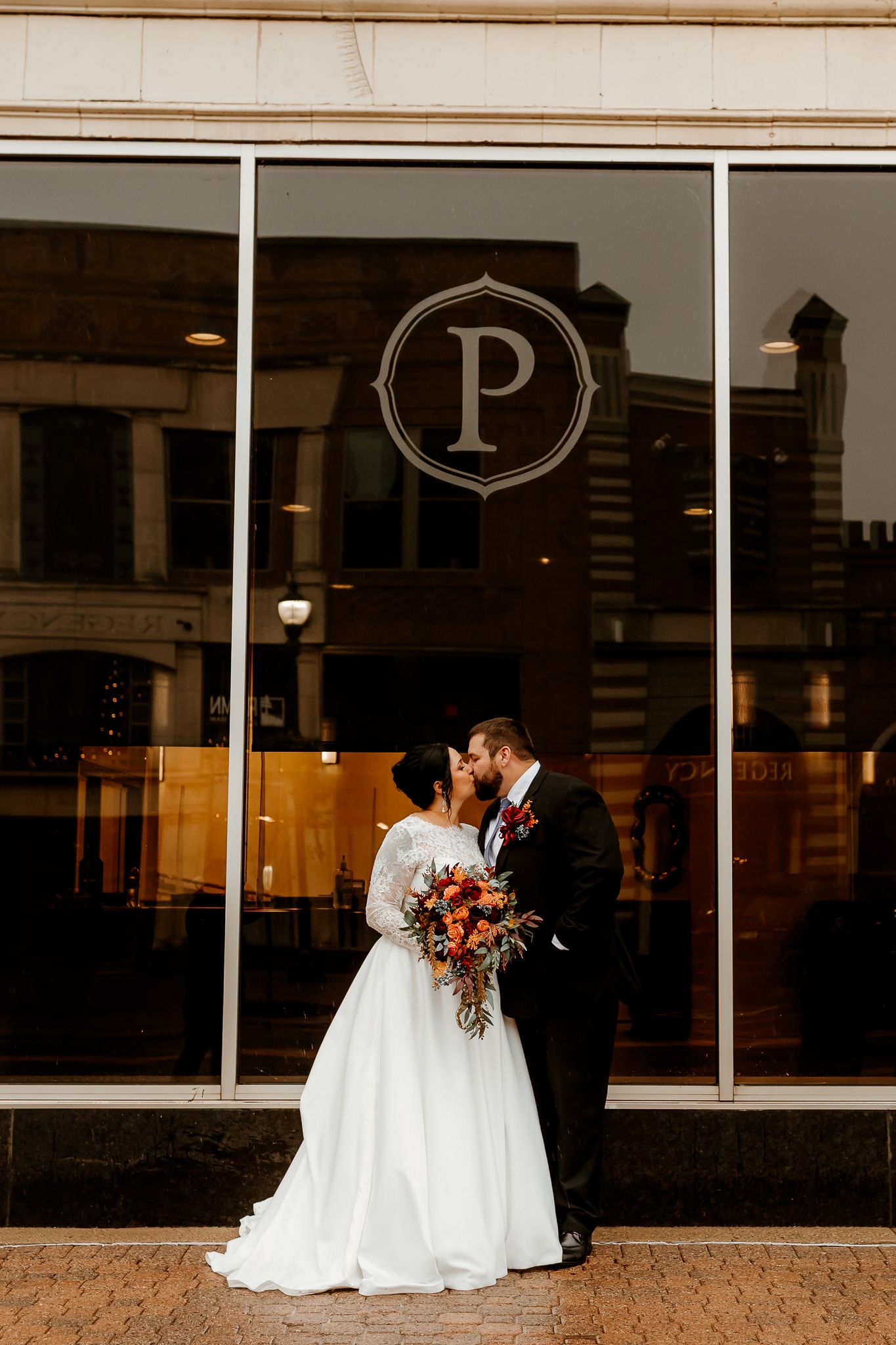 Bride in a white lace gown and groom in a dark suit kiss while holding an orange bouquet in front of large windows with a circular P logo.