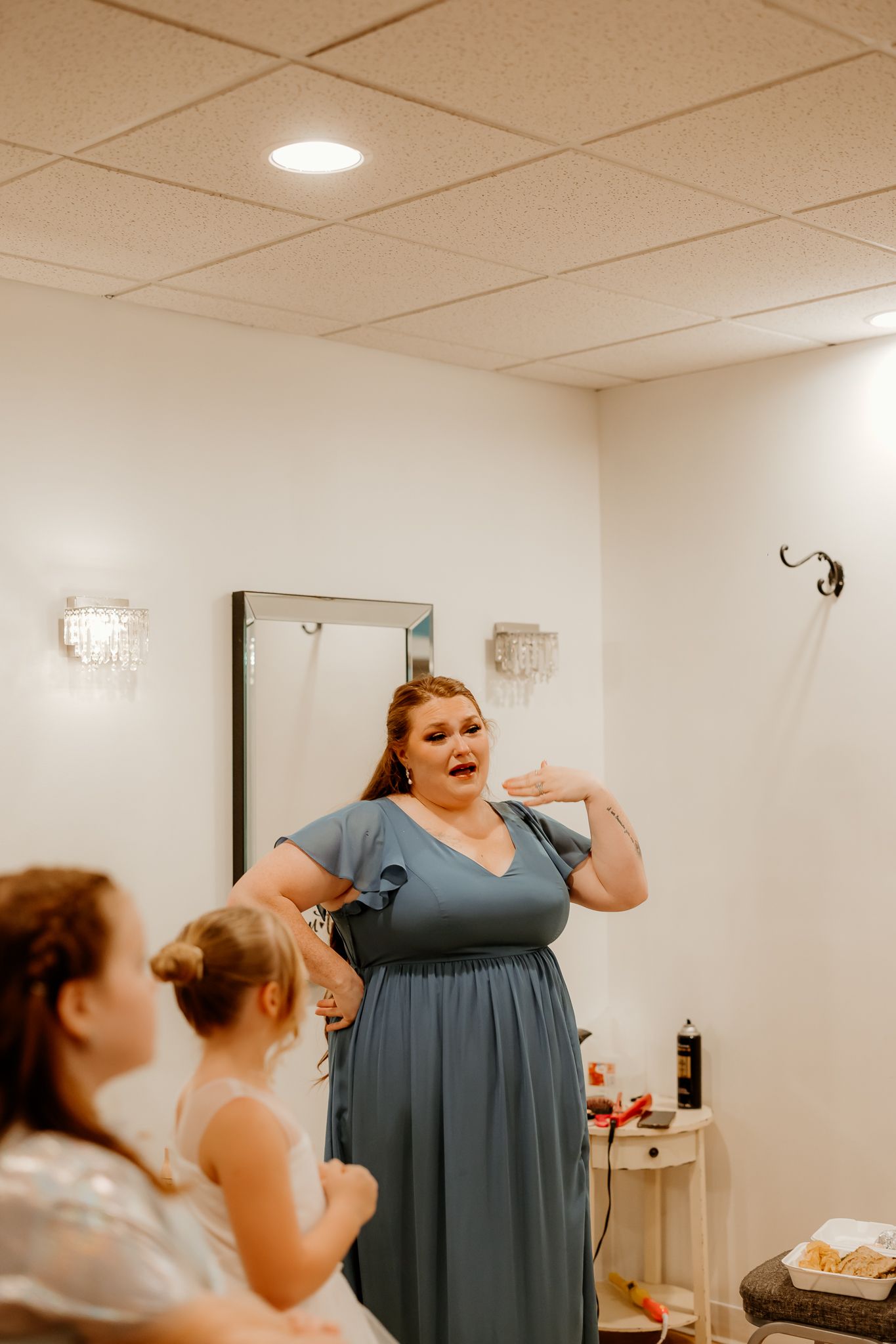 Woman in a dusty blue dress gestures expressively while two young girls watch in a bright getting-ready room with a mirror and wall sconces.