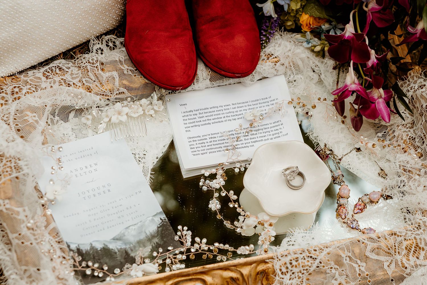 Elegant wedding flat lay with red boots, floral invitations, diamond ring, pearl details, and colorful bouquet on lace fabric.