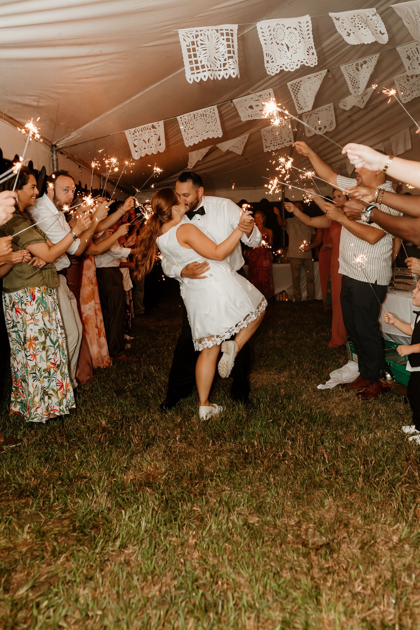 Bride and groom share a romantic kiss during a sparkler send-off under a tent adorned with papel picado at their wedding reception.