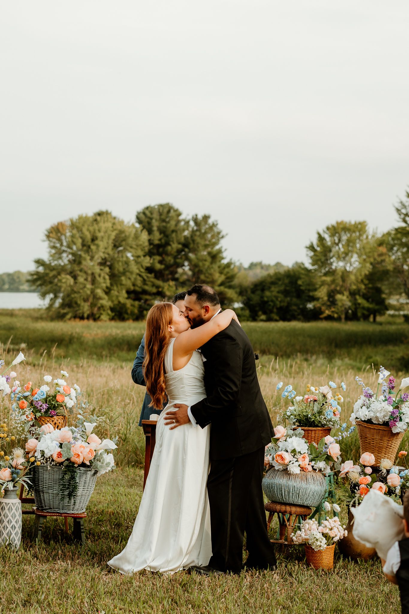 Bride and groom share first kiss outdoors at rustic field wedding ceremony surrounded by colorful floral arrangements in vintage pots.