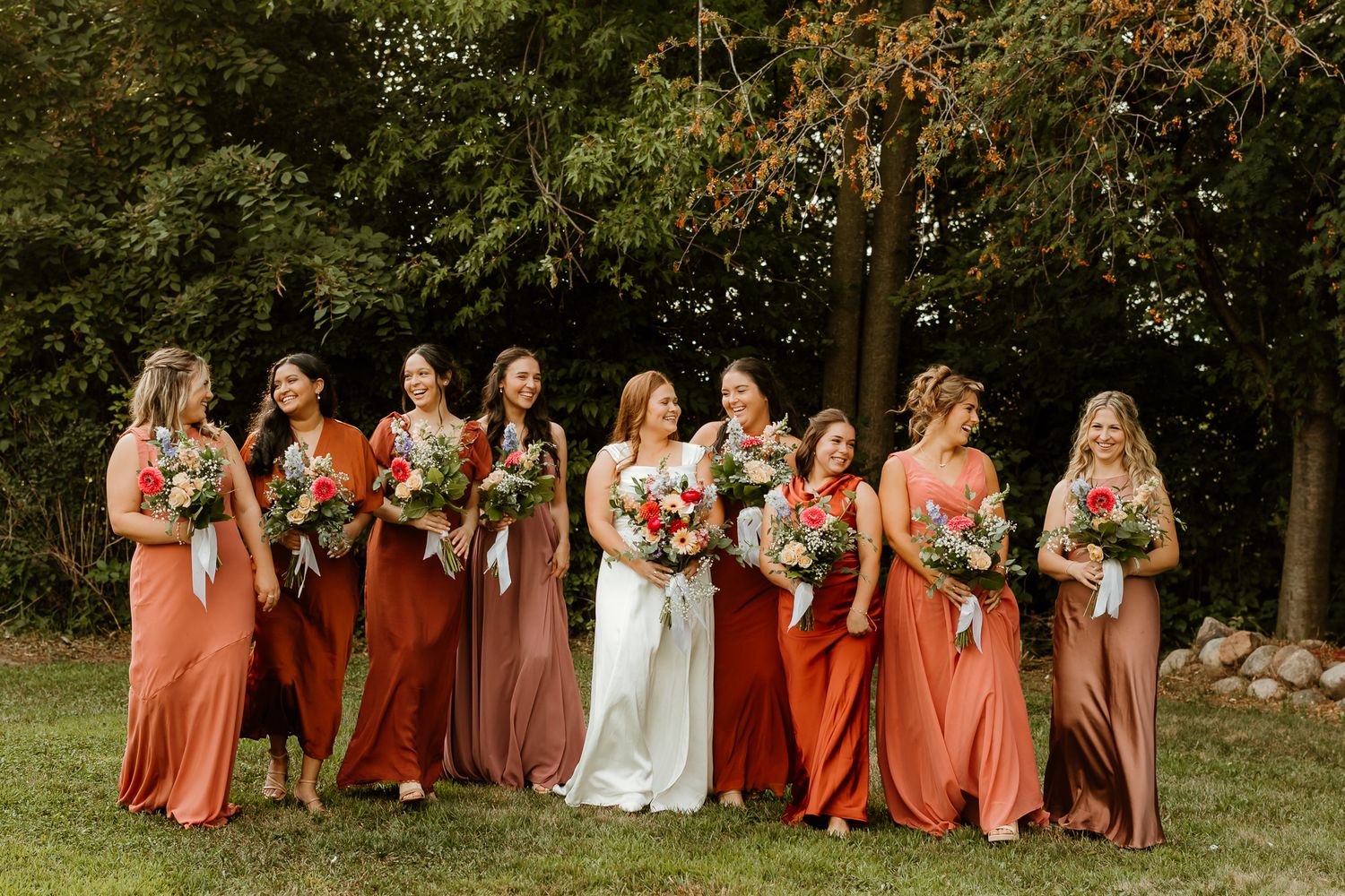 Bride and bridesmaids in rust and terracotta dresses holding colorful bouquets outdoors at a fall wedding.