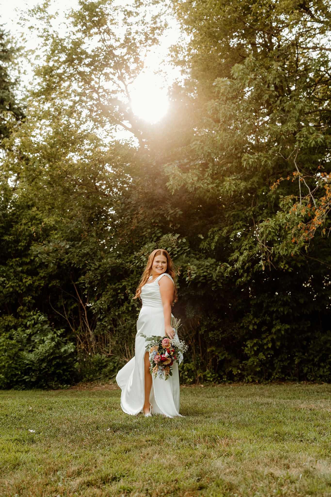 Bride in white gown holding bouquet stands in sunlit garden, golden hour light filtering through lush green trees.