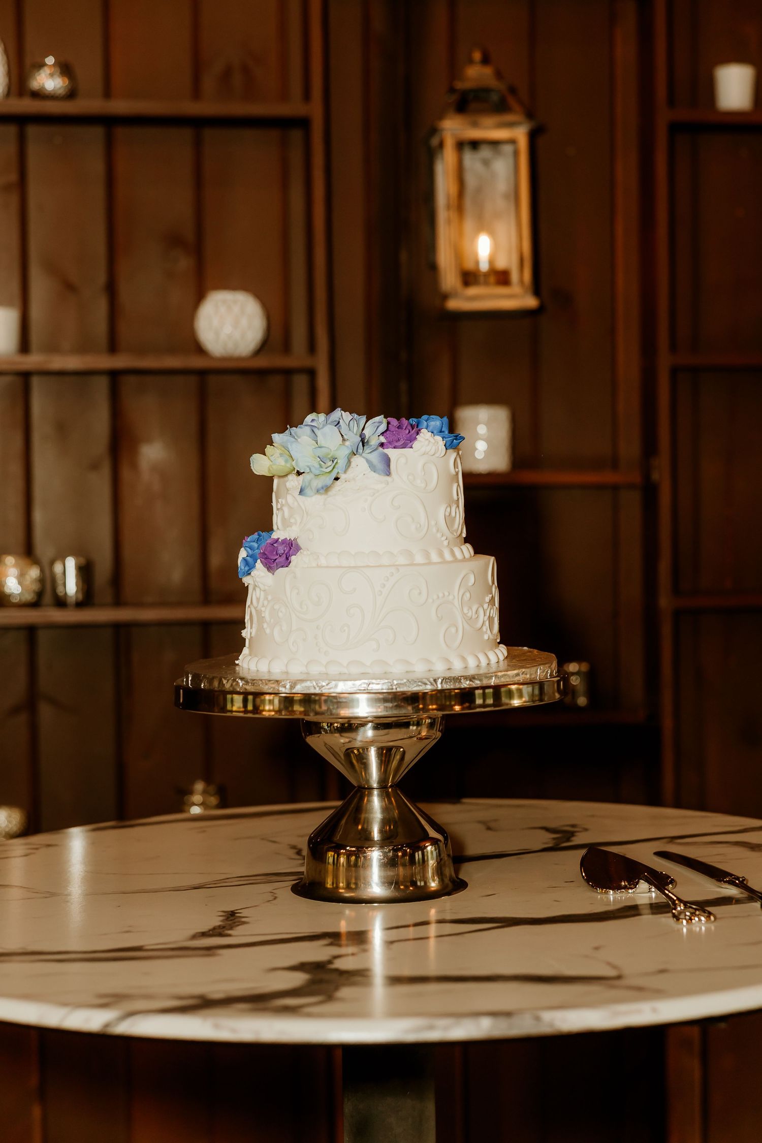 Two-tier white wedding cake with blue and purple floral decorations on a silver stand, displayed on a marble table.