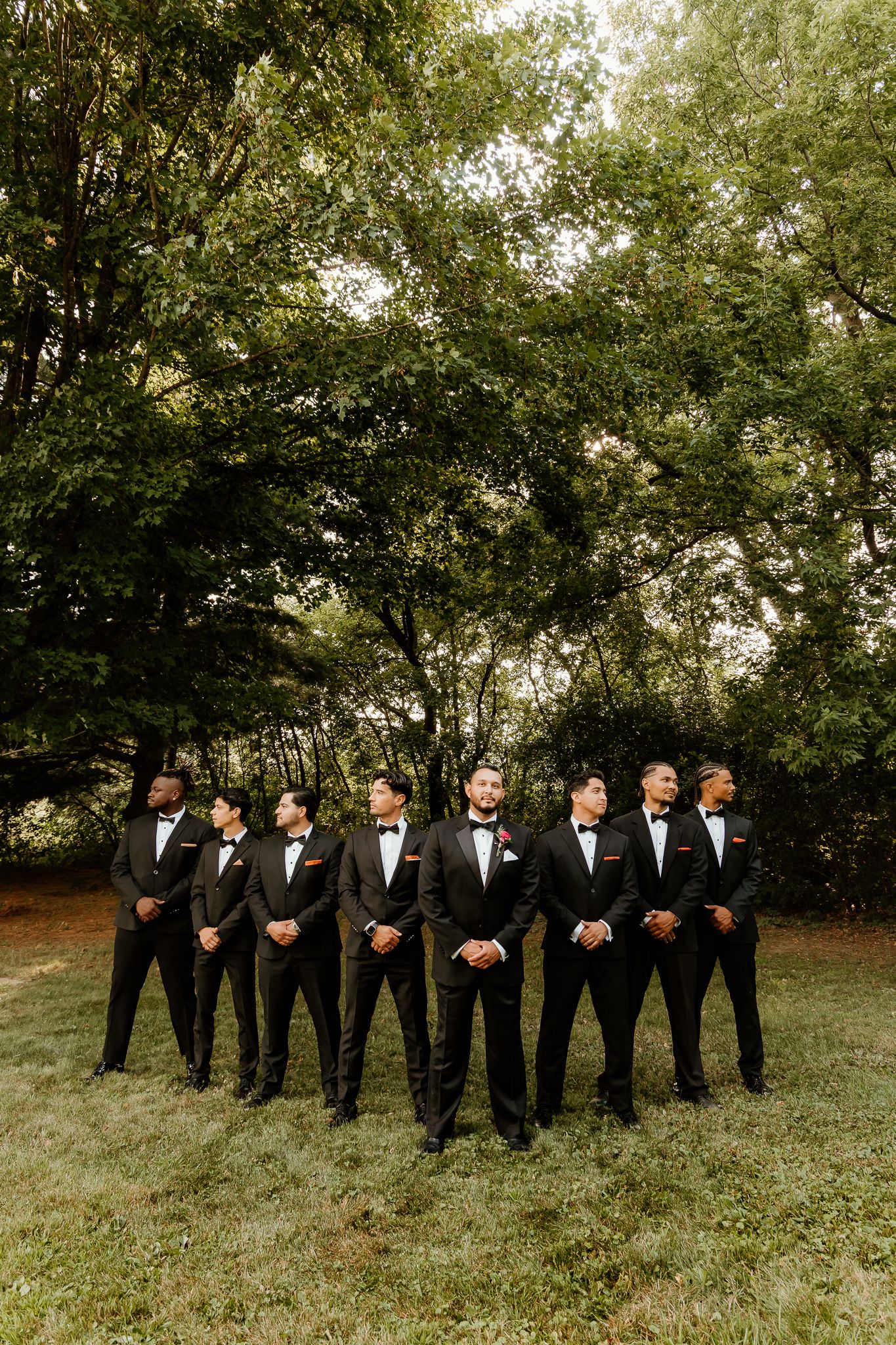Seven groomsmen in black tuxedos and bow ties pose outdoors on grass with lush green trees in the background.