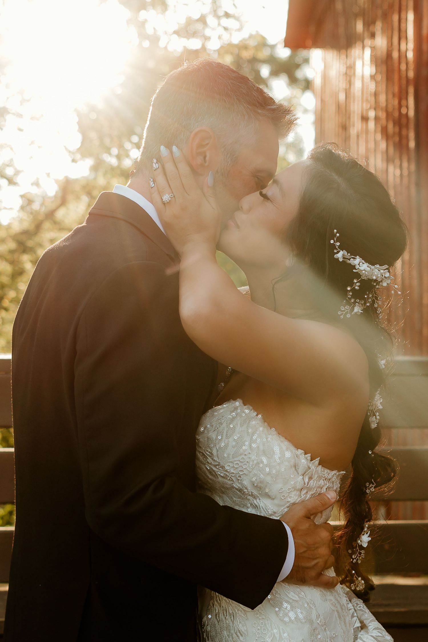 A bride and groom share a romantic kiss bathed in golden sunlight, she in lace gown with floral hair piece.