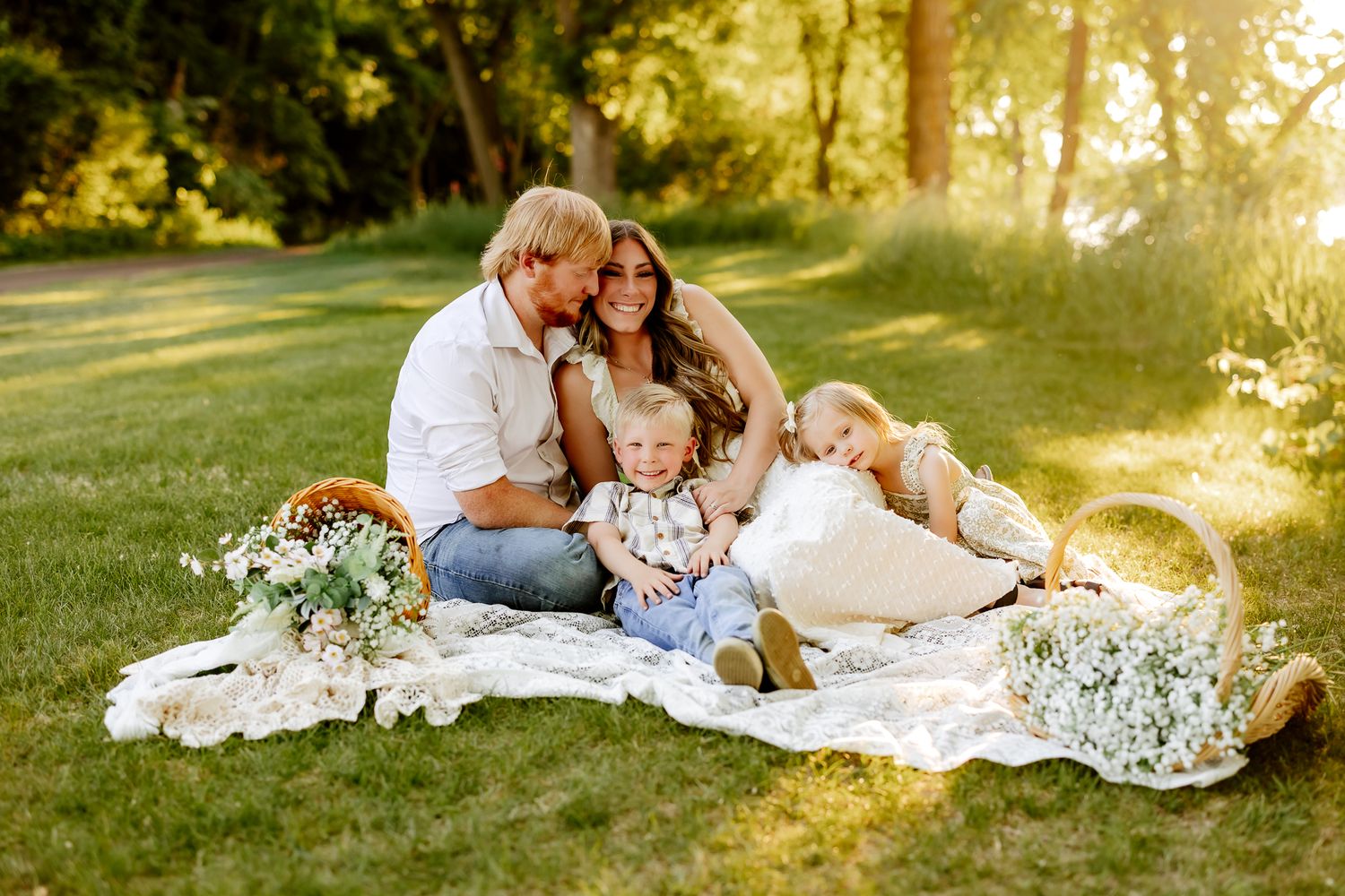 A happy family of four enjoys a sunny outdoor picnic on a white blanket surrounded by flower baskets.