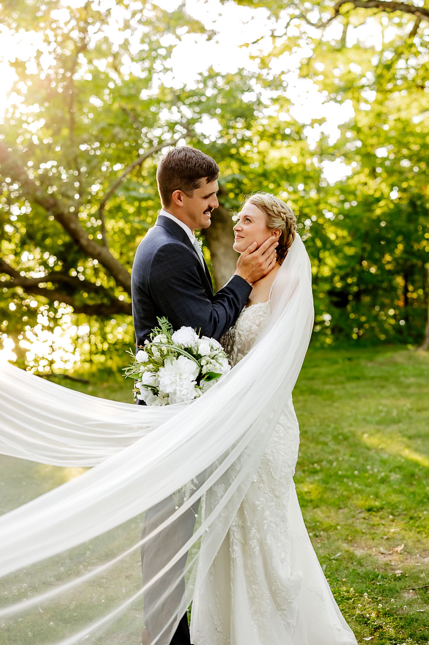 Bride and groom embrace outdoors, her veil flowing in the breeze, surrounded by lush green trees in golden sunlight.