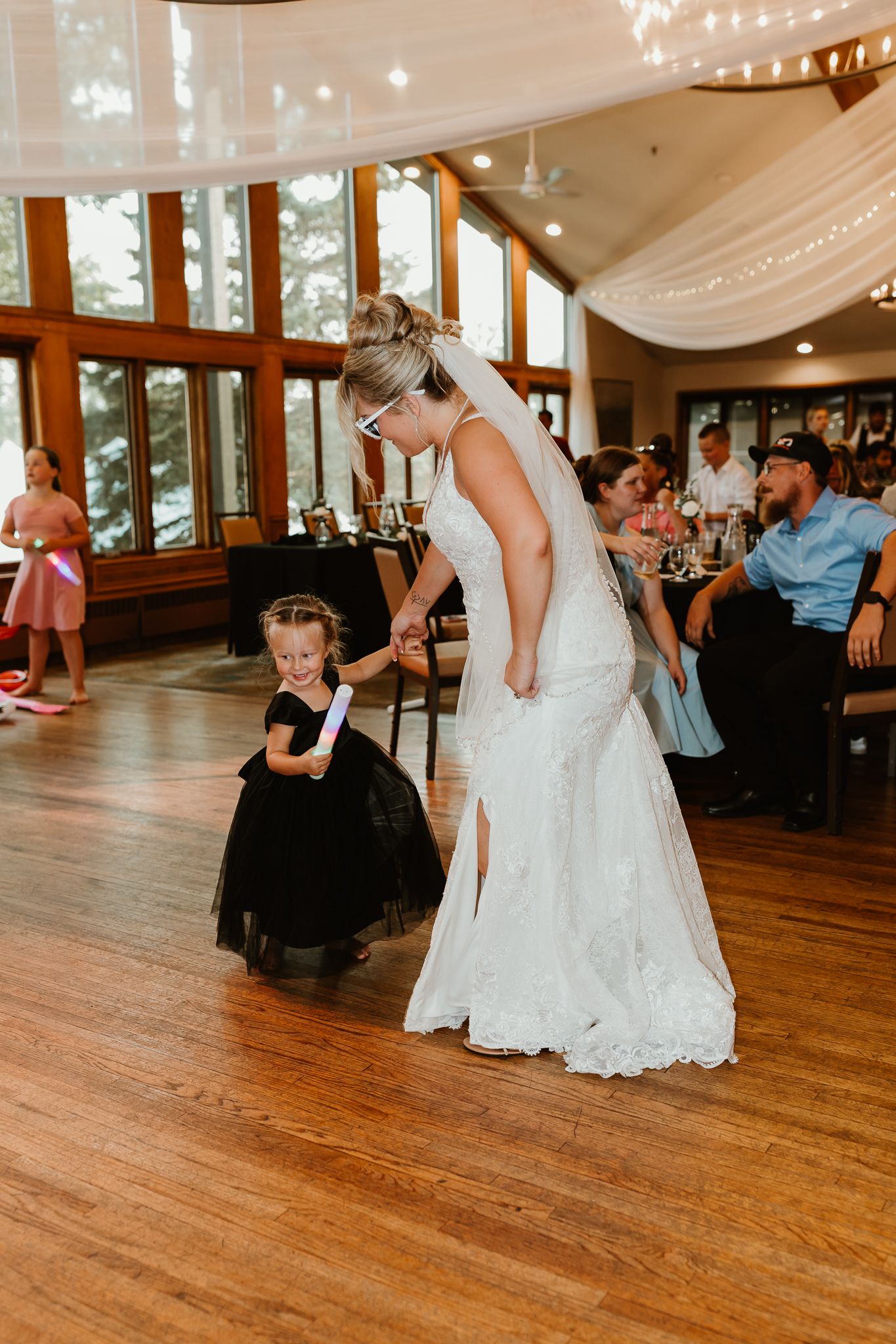 Bride in white lace gown dances with young flower girl in black dress at wedding reception.