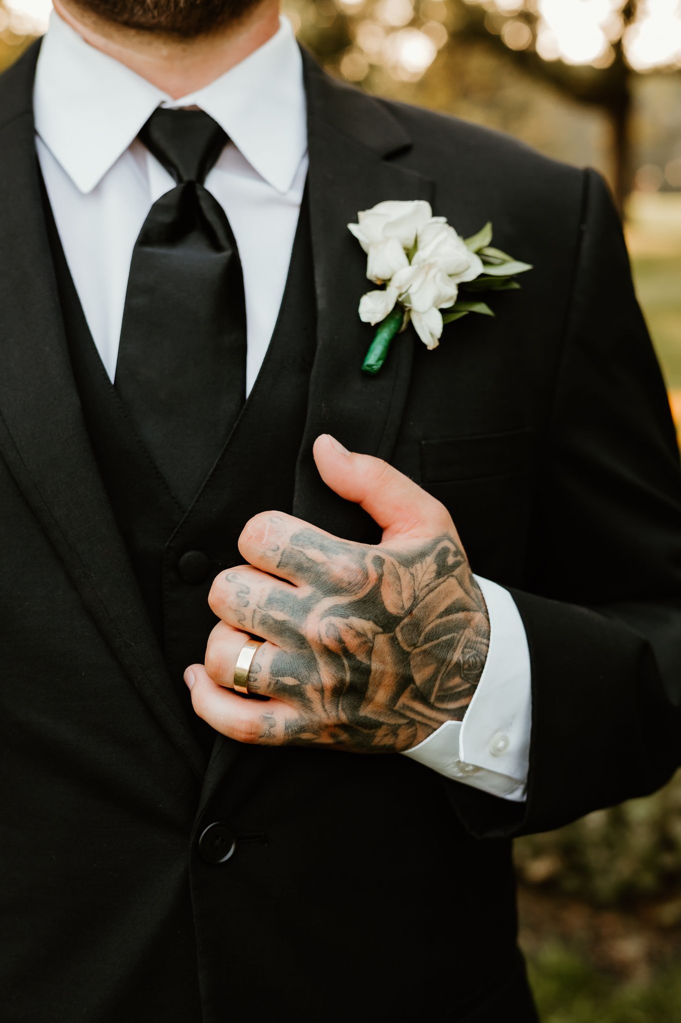 Groom in black suit with white boutonniere and tattooed hand wearing gold wedding band.
