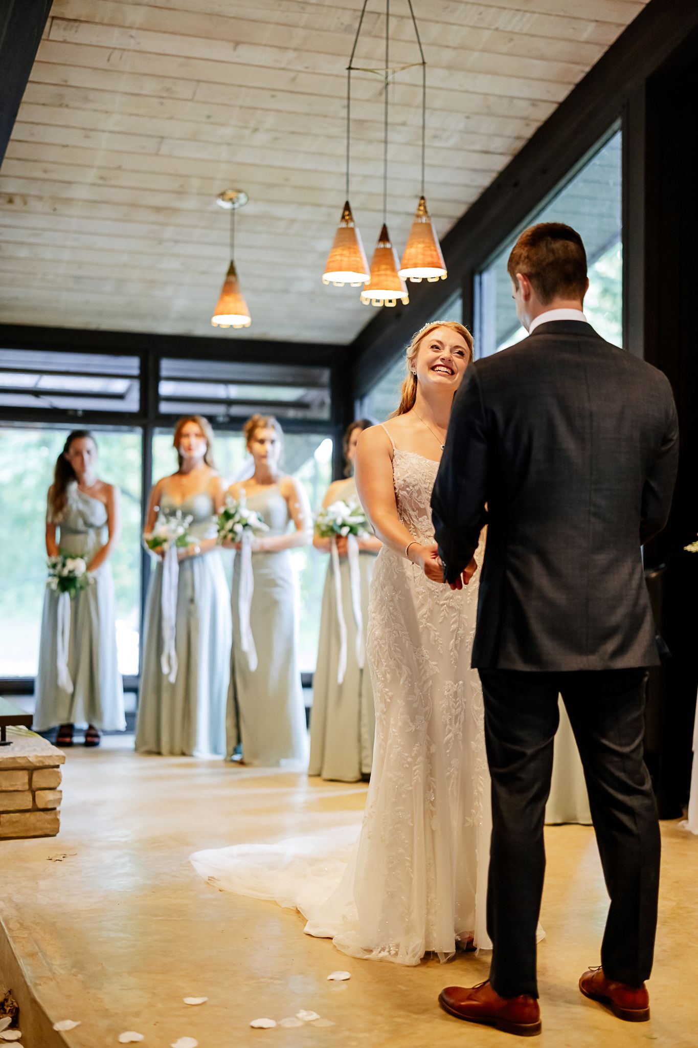 Bride and groom hold hands at indoor wedding ceremony with bridesmaids in sage green gowns holding white bouquets.