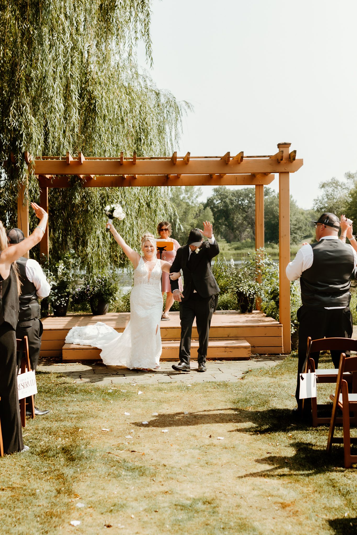 Joyful newlyweds celebrate after outdoor wedding ceremony under wooden pergola surrounded by guests and weeping willow tree.