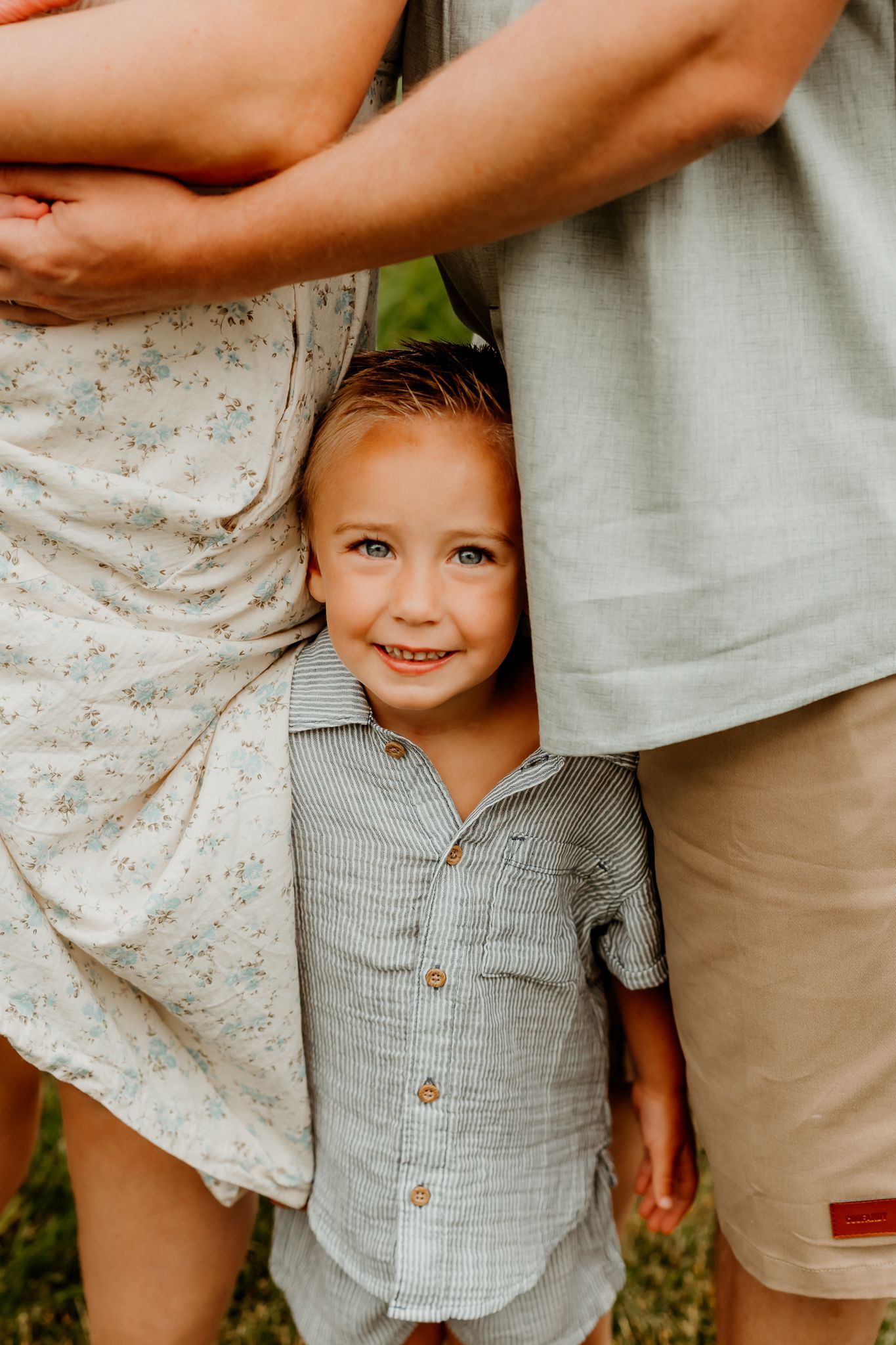 A smiling young boy in a gray button-up shirt stands between two adults during a family photo session outdoors.