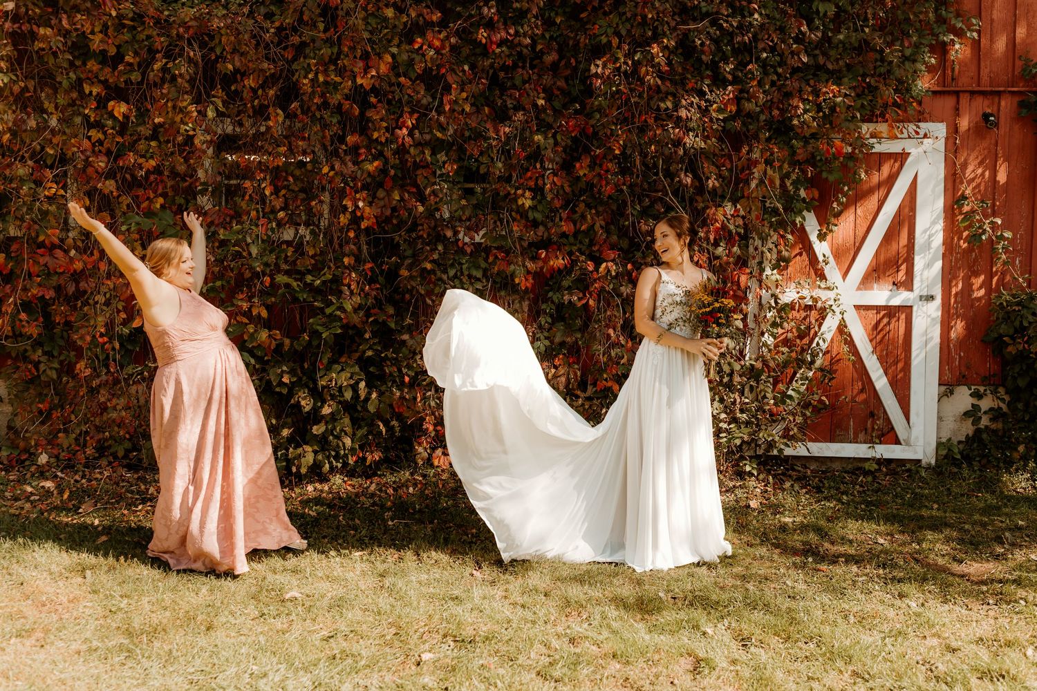 Bridesmaid in a blush pink dress lifts her arms as a bride in a white gown stands holding a bouquet near a rustic red barn door.