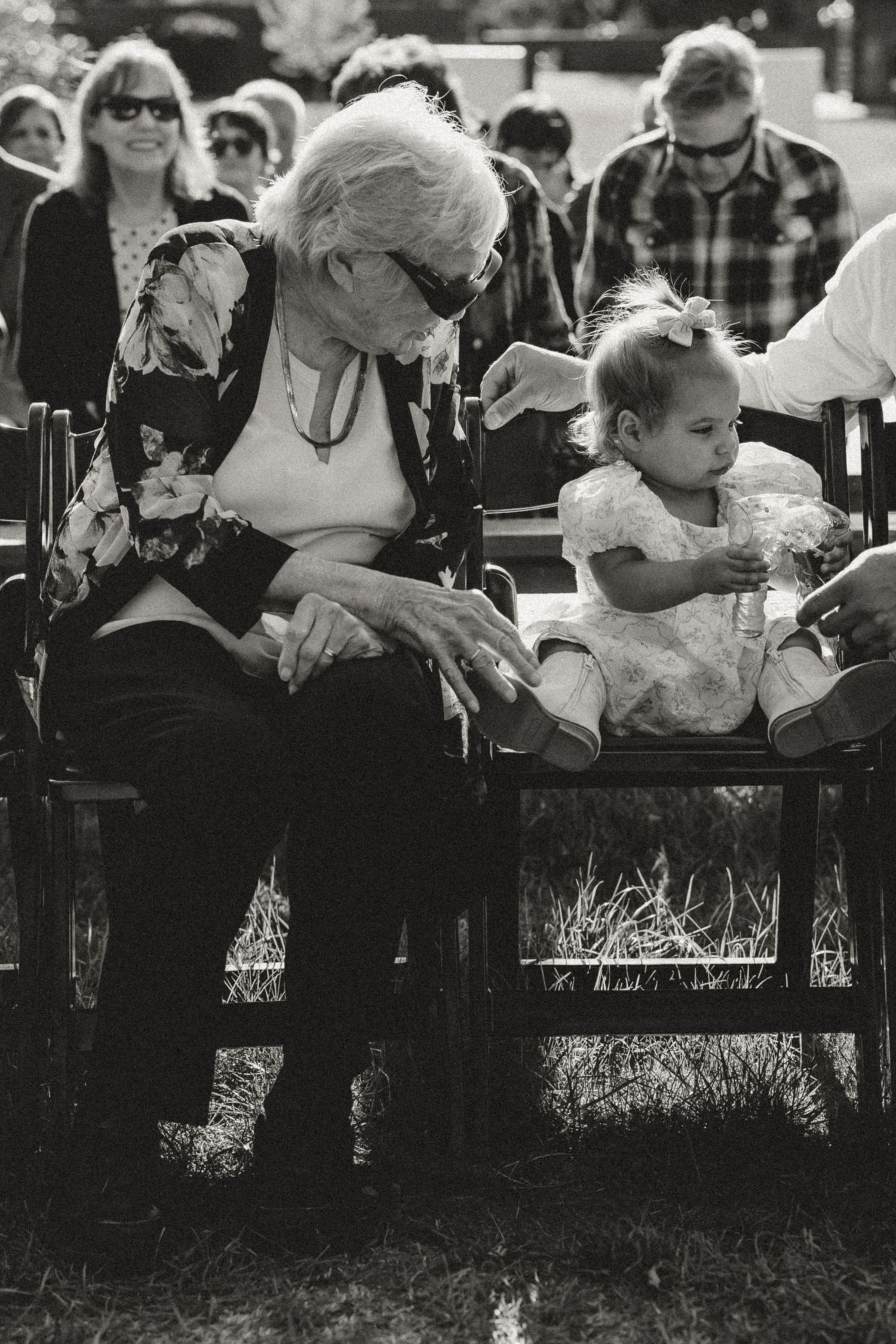 Elderly woman in sunglasses and a floral jacket sits beside a toddler in a white dress and bow on wooden chairs at an outdoor gathering.