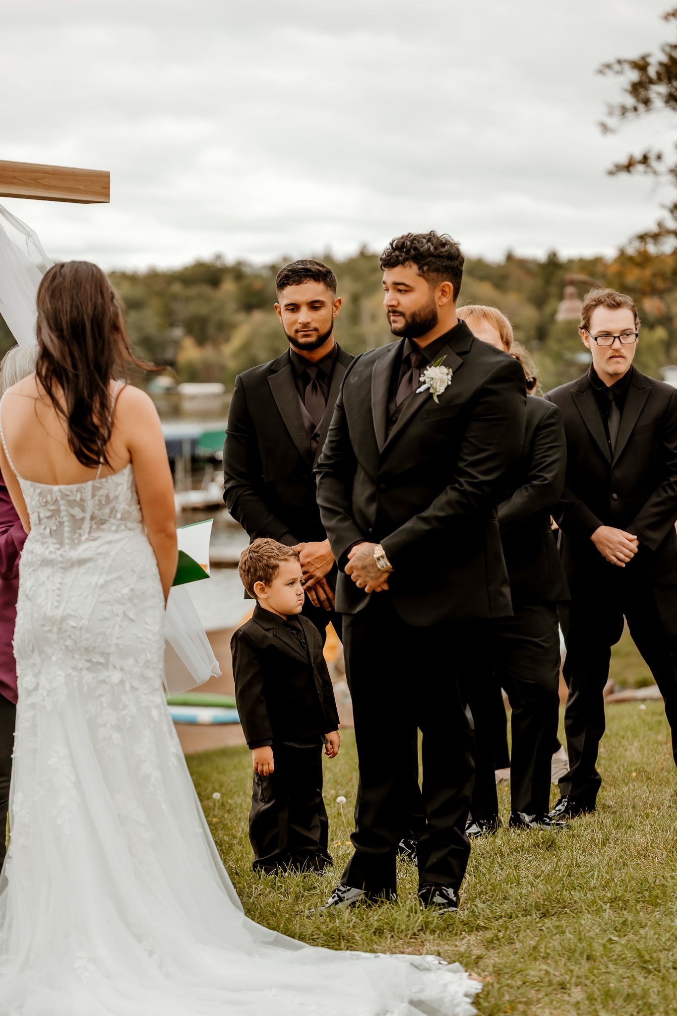 Groom in a black suit stands with groomsmen and a young ring bearer as a bride in a lace gown faces them at an outdoor wedding ceremony.
