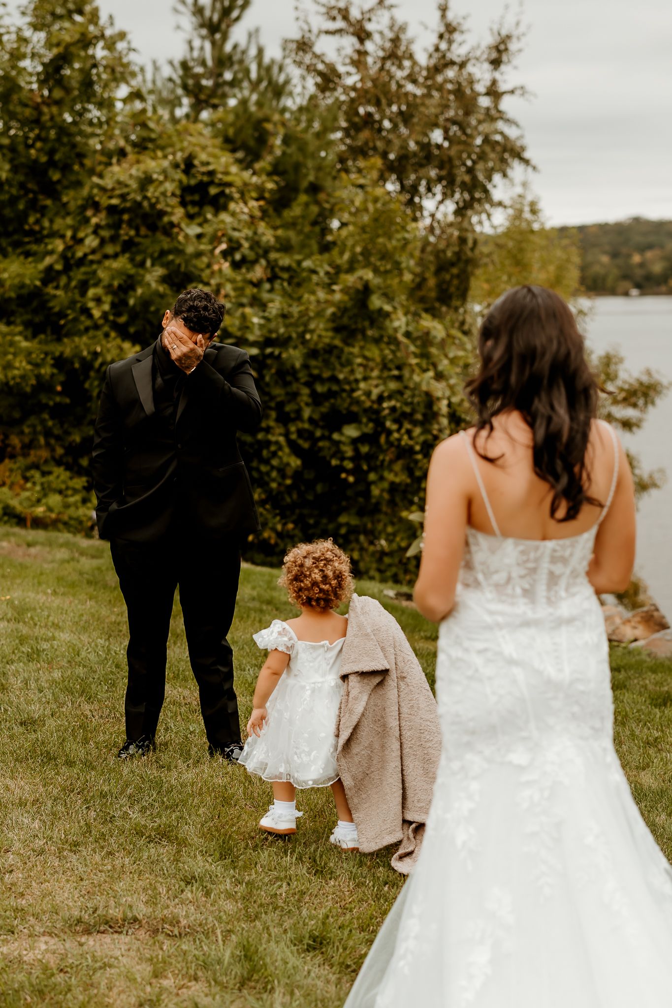 Emotional groom covers his face during a first look as a bride in a lace gown and a toddler in white stand on a grassy lakeside lawn.
