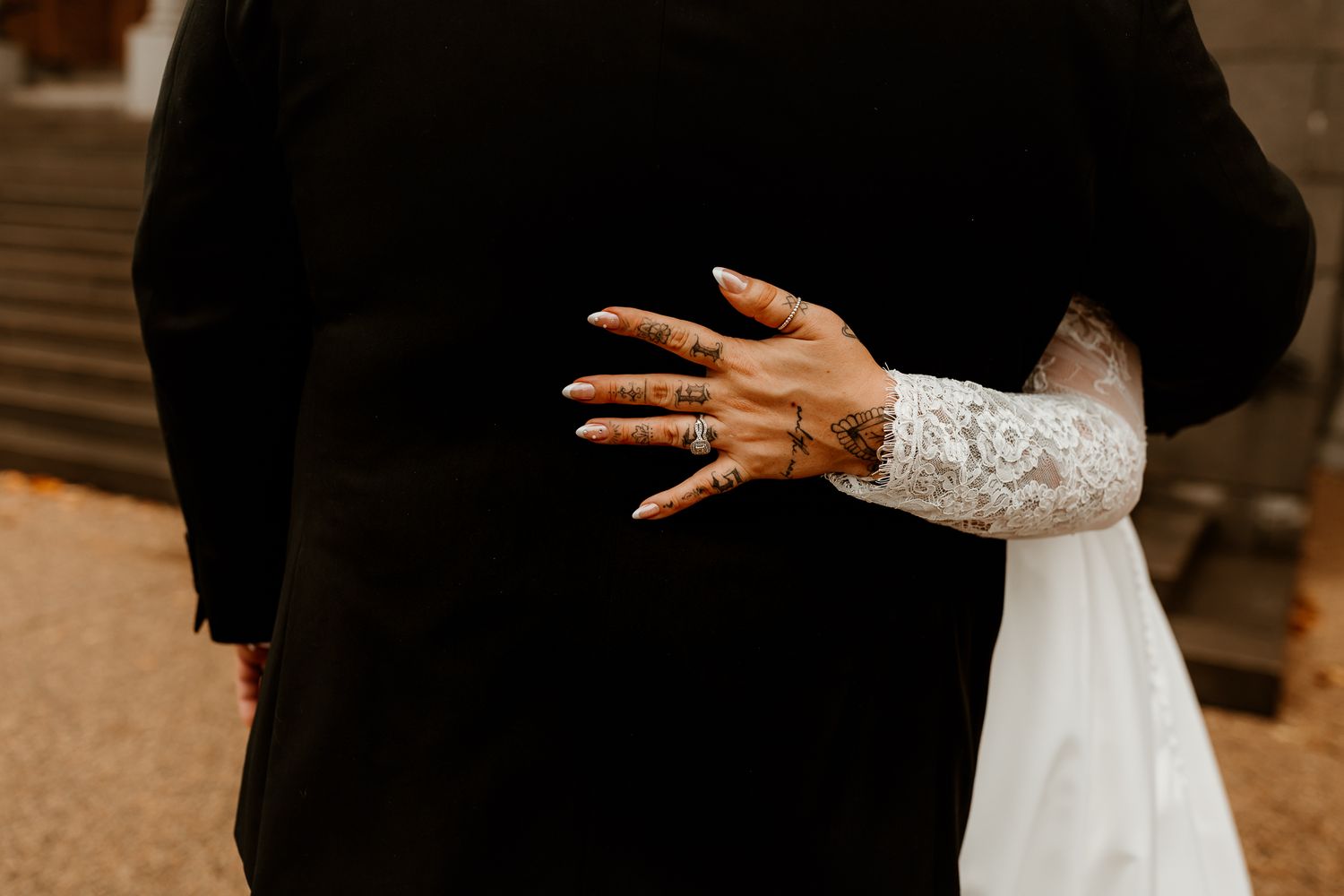 Bride in a white lace-sleeved gown rests her hand with henna-style tattoos and pink nails on groom's black suit jacket during an embrace.