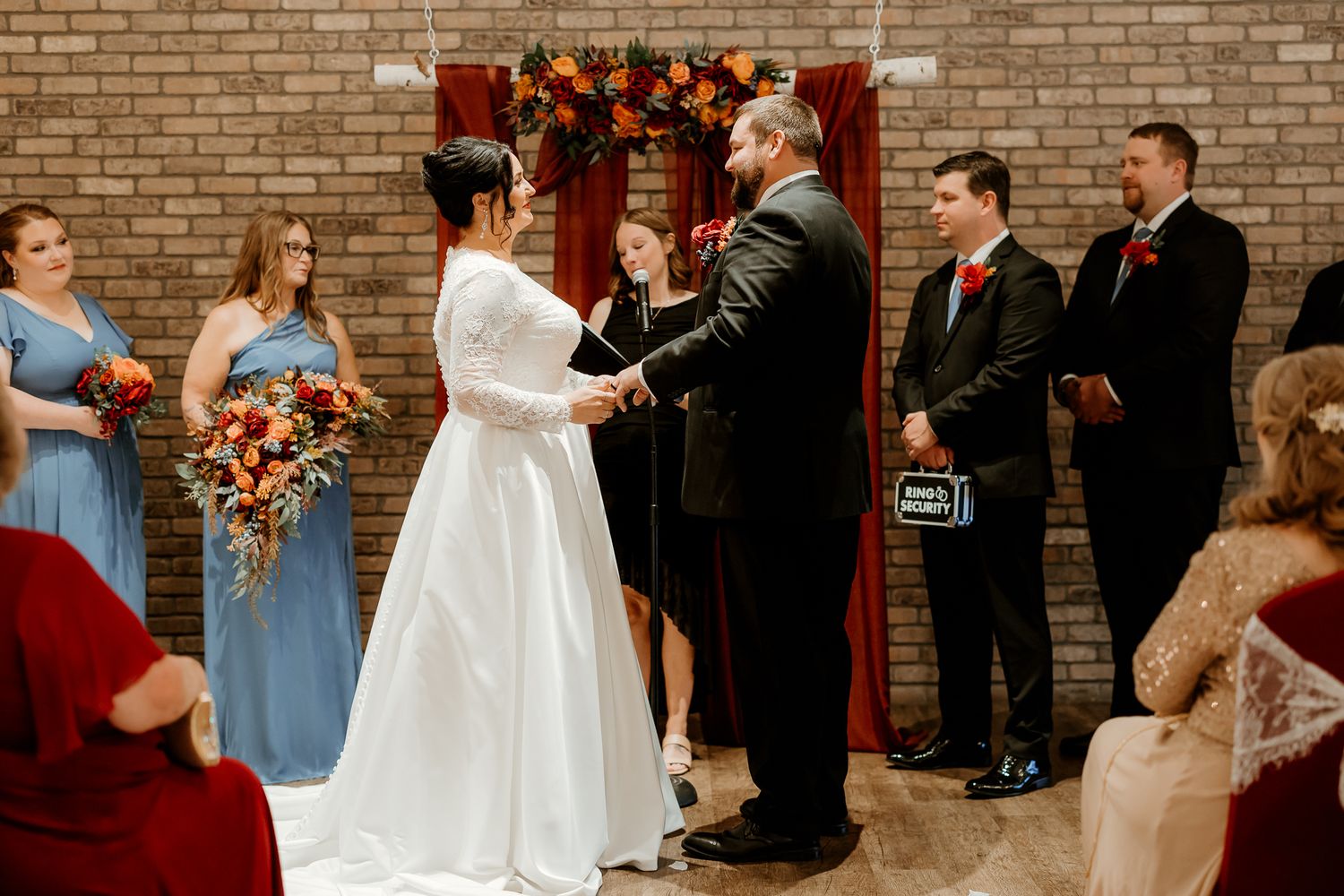 Bride in white gown and groom in dark suit exchange vows at an indoor wedding ceremony as bridesmaids, groomsmen and guests look on.