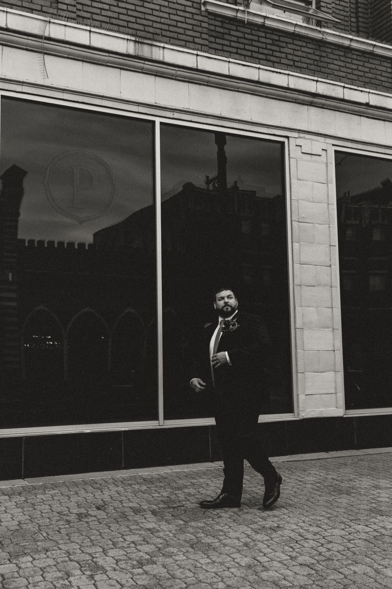 Bearded groom in a dark suit and boutonniere walks past a large storefront window reflecting historic buildings in black and white.