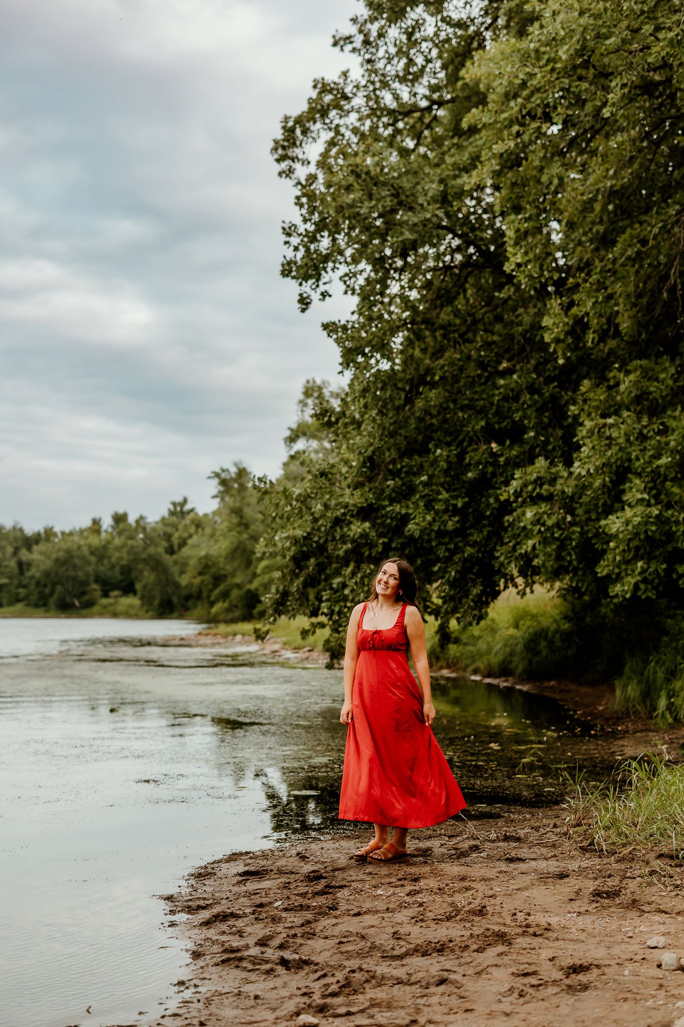 Woman in red dress standing on riverbank with lush green trees and cloudy sky backdrop.