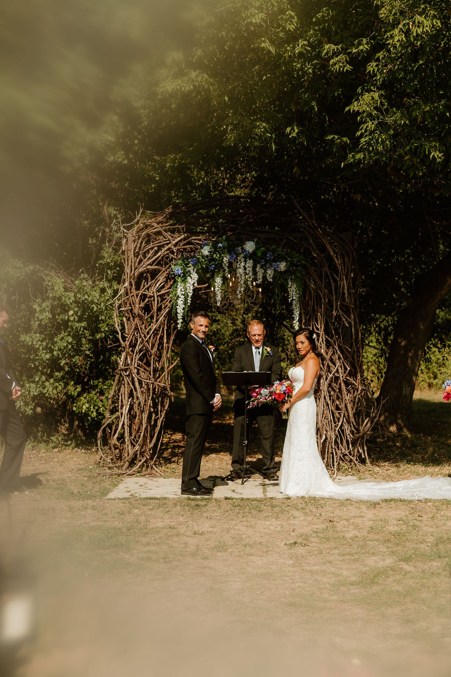 Bride and groom exchange vows under a rustic twig arch surrounded by lush green trees during an outdoor wedding ceremony.