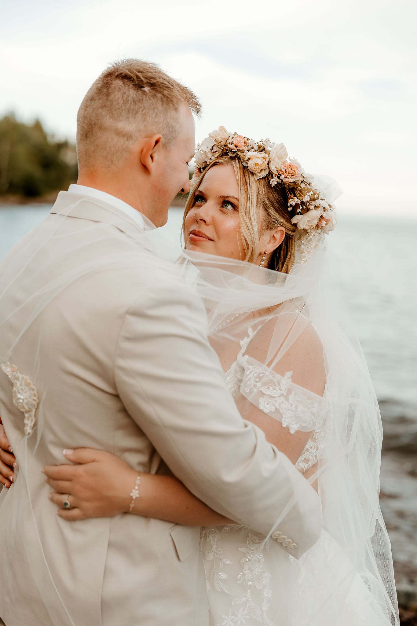 Bride in floral crown and lace veil embraces groom in beige suit by the water at their beach wedding.