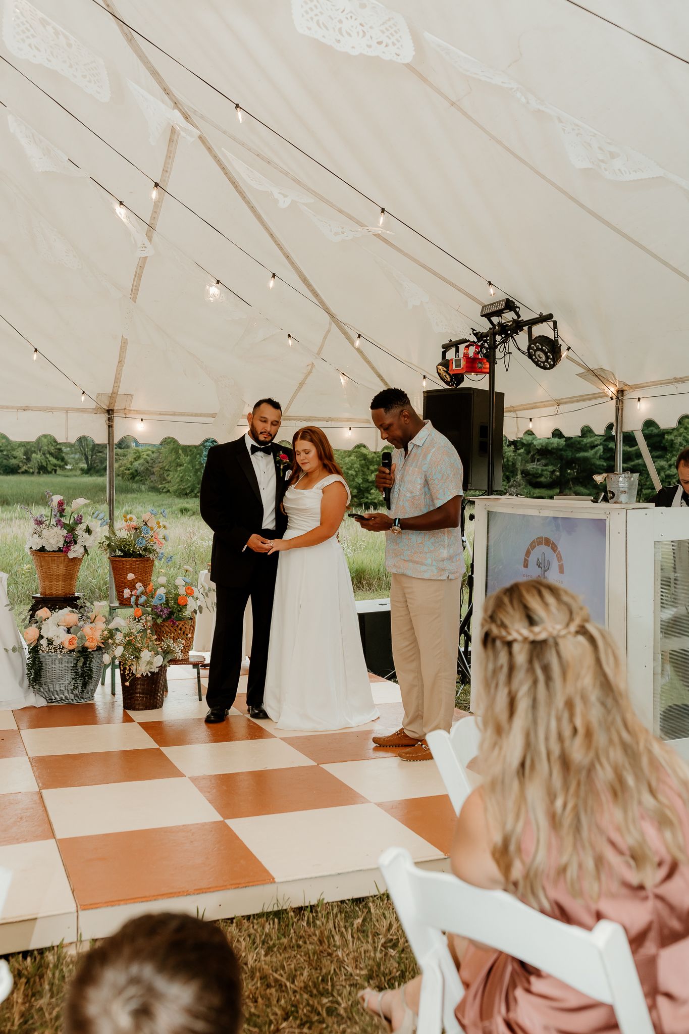 Bride and groom exchange vows under a white tent with string lights on a checkered dance floor at an outdoor wedding.