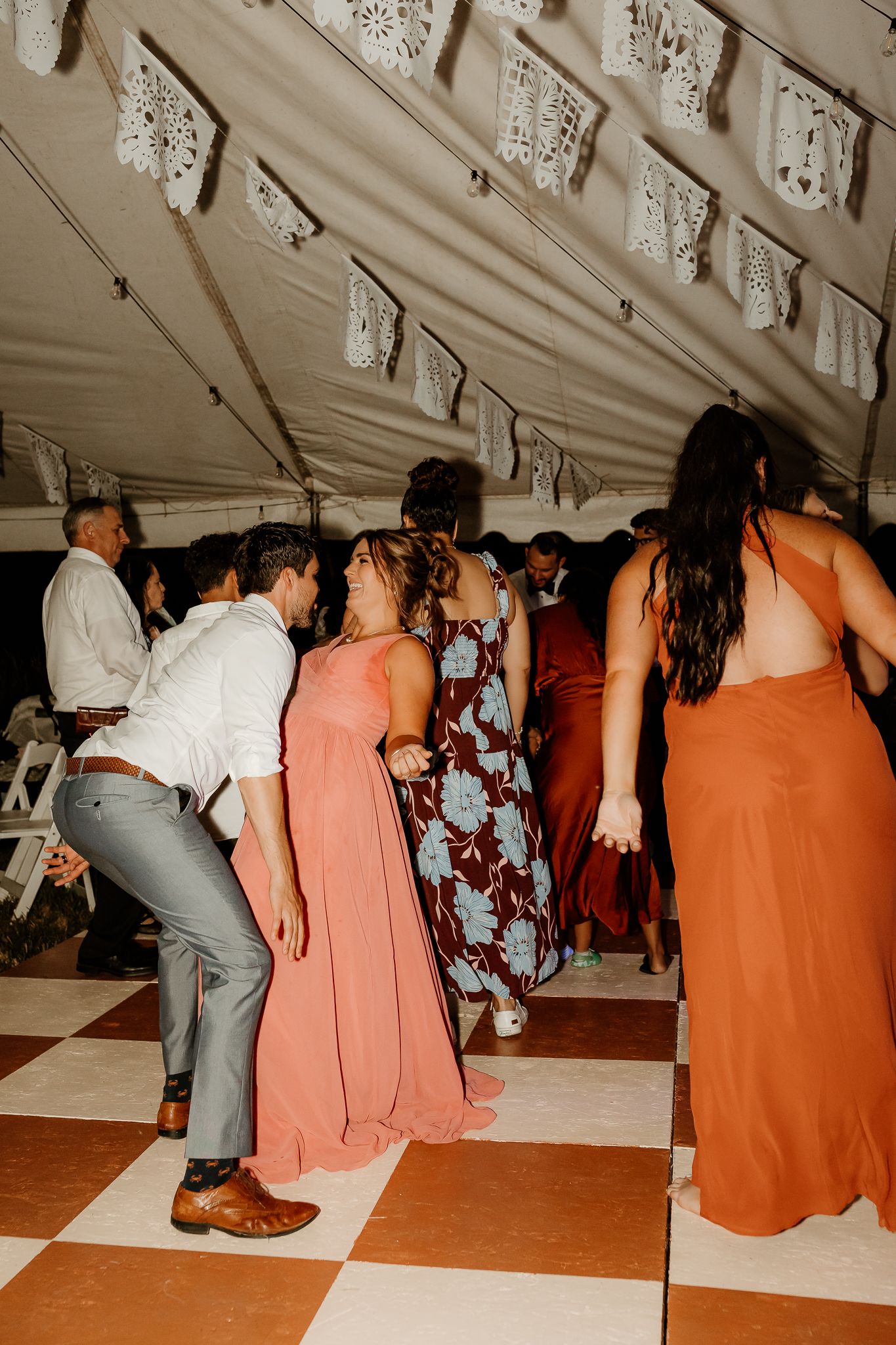 Guests dancing at a wedding reception under a tent with papel picado decorations and checkered dance floor.